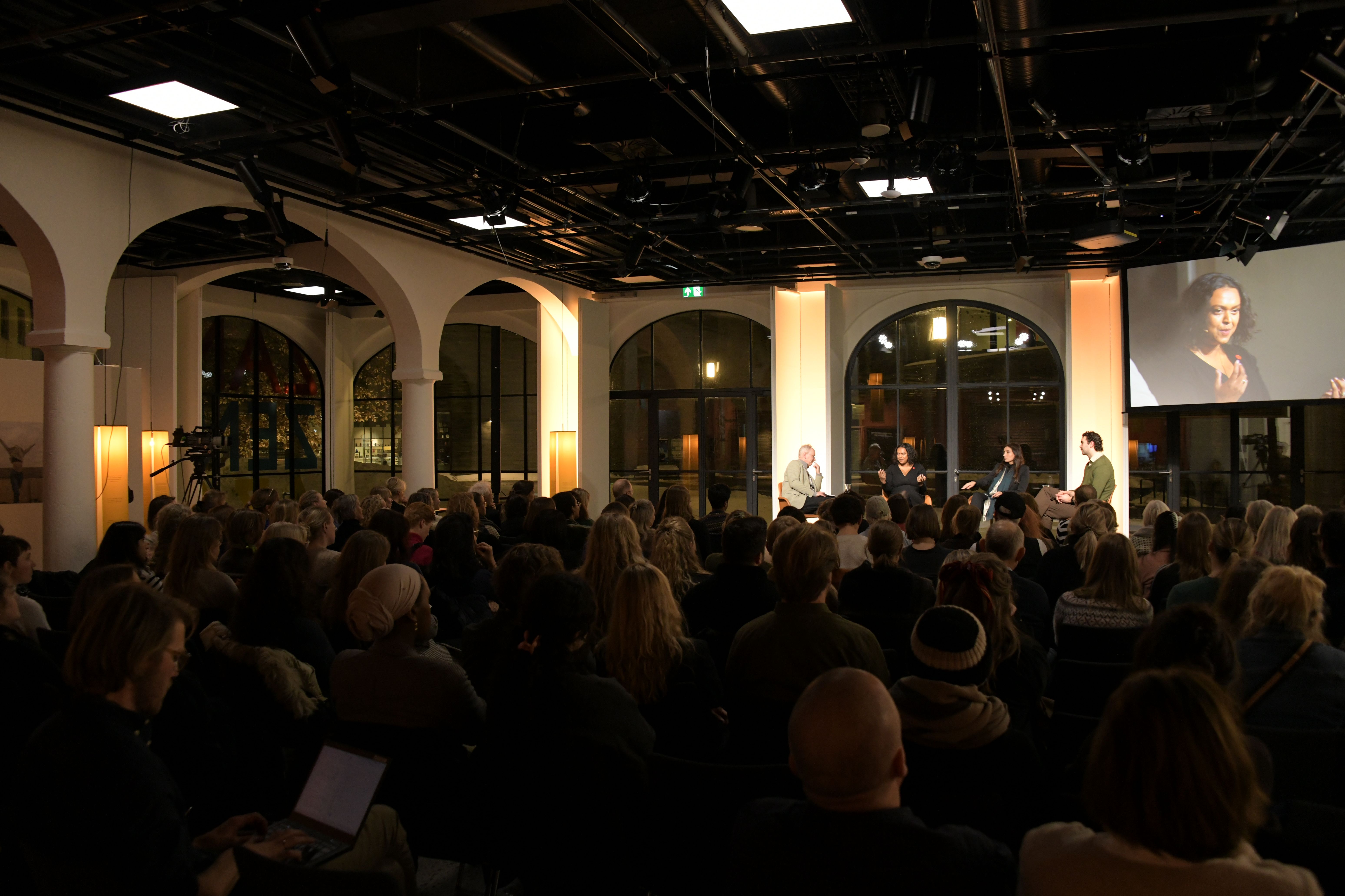 her er toghallen med et arrangement og scene på kveldstid, og en stor skjerm som viser talerne på scenen.