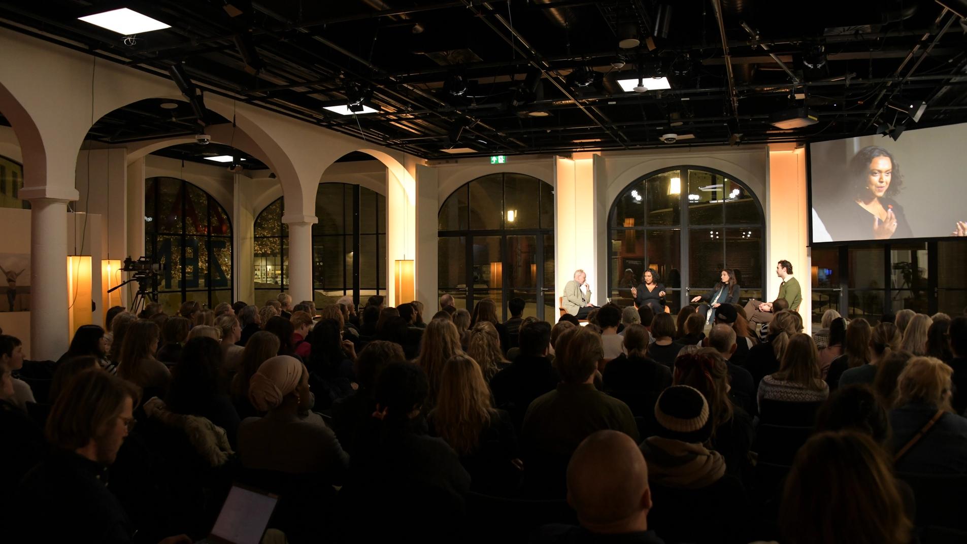her er toghallen med et arrangement og scene på kveldstid, og en stor skjerm som viser talerne på scenen.
