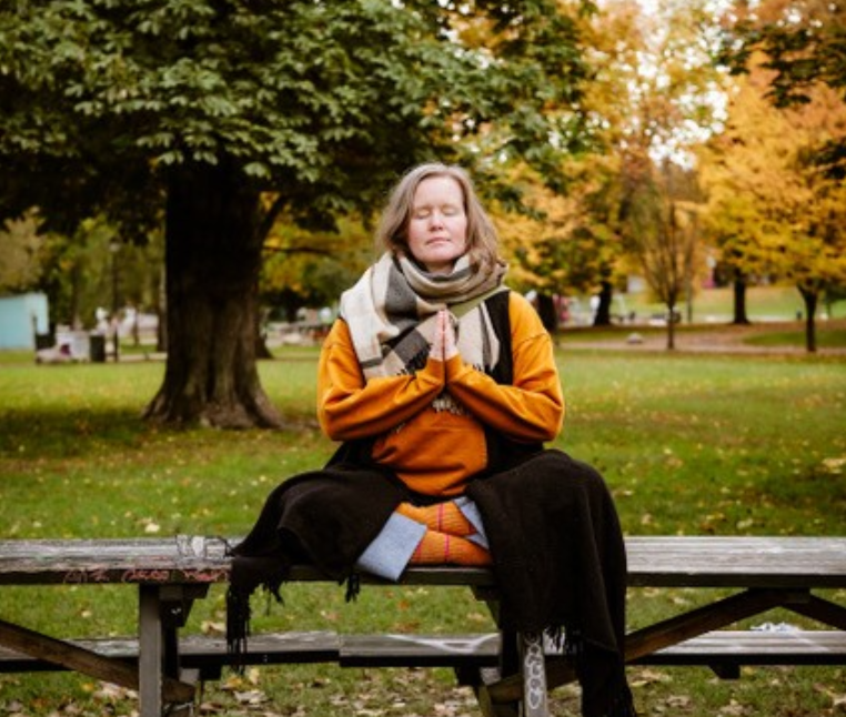 Karina som sitter i lotus posisjon på en benk ute i en park.
