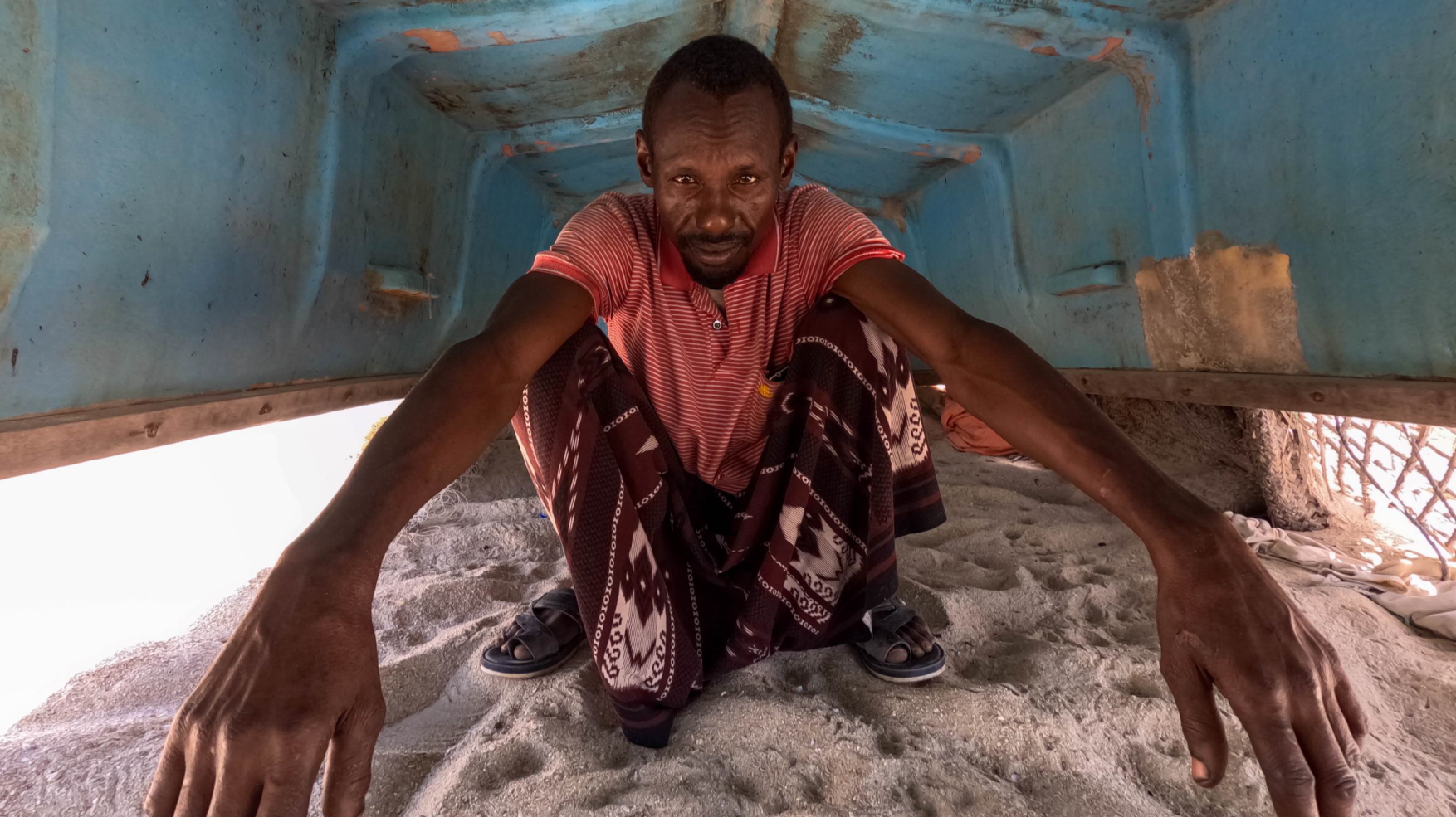 En mann som sitter under en oppned båt på sand.