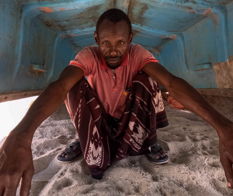 En mann som sitter under en oppned båt på sand.