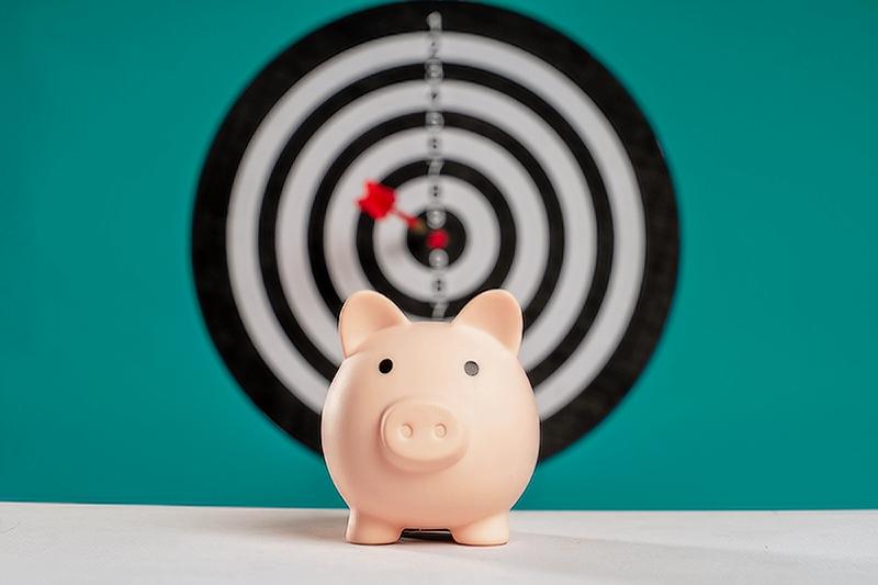 An image of a piggy bank with a dart board with a dart in bullseye in the background