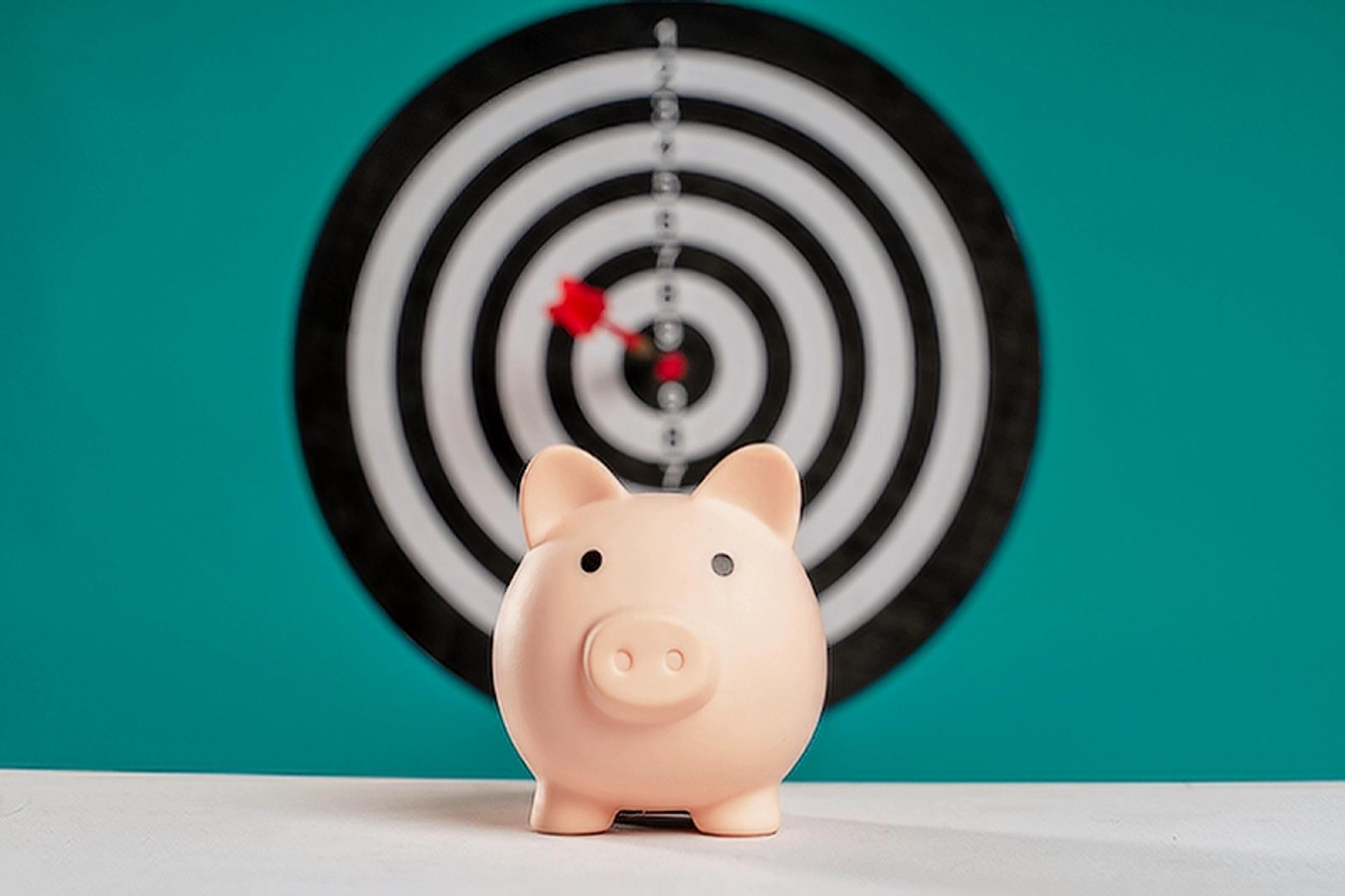 An image of a piggy bank with a dart board with a dart in bullseye in the background