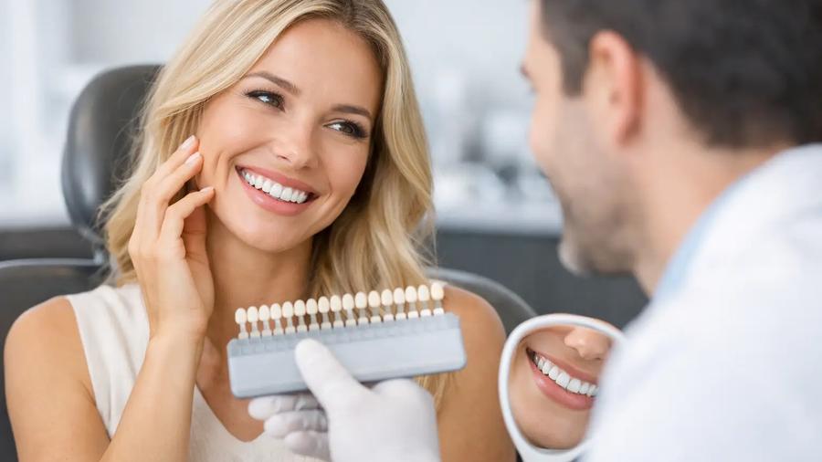 Dentist holding a veneer shade guide and mirror up to a smiling patient during a cosmetic dental consultation.