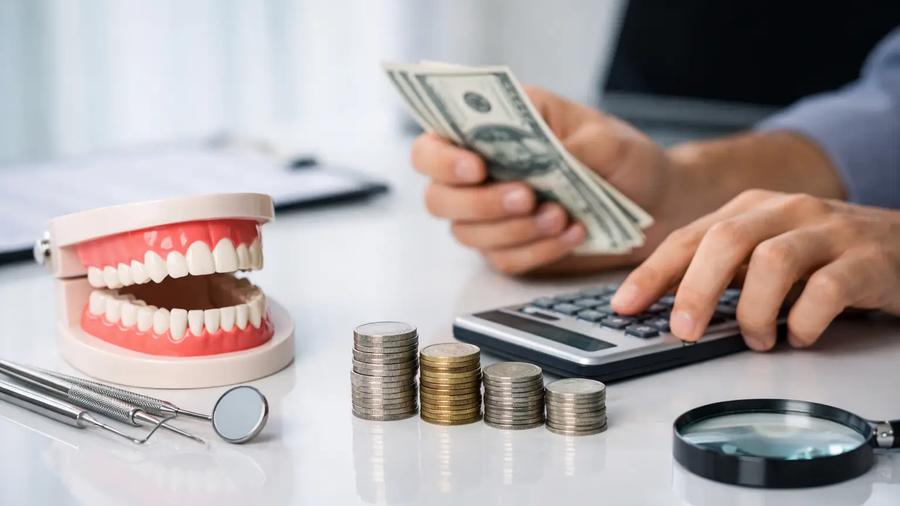 Hands counting cash and using a calculator beside a dental jaw model, stacked coins, tools, and a magnifying glass on a desk.