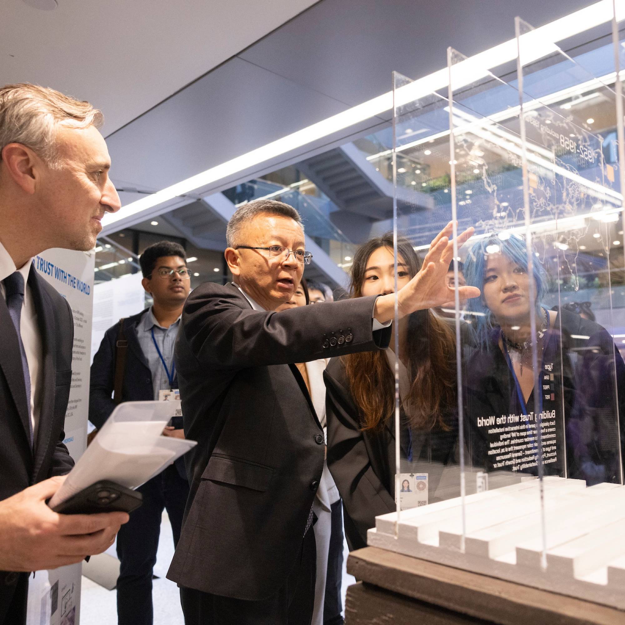 Bo Li speaking in front of a glass exhibit with several people looking at the exhibit.