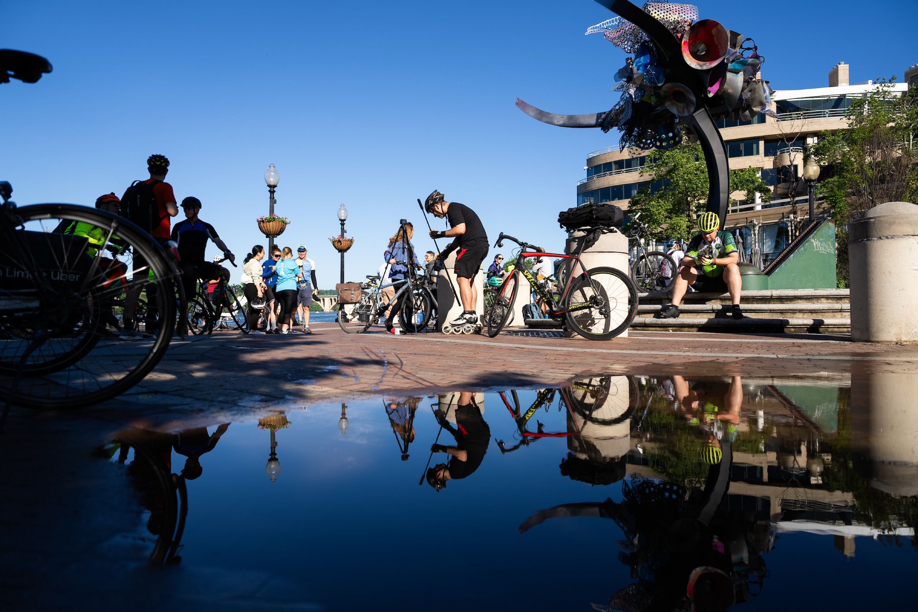 A puddle in the foreground reflecting a daytime scene with bikes, a group of people and a man on rollerblades.