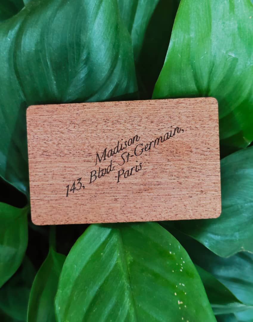 Carte-clé en bois gravée « Madison, 143 Bd St-Germain, Paris », posée sur des feuilles vertes
