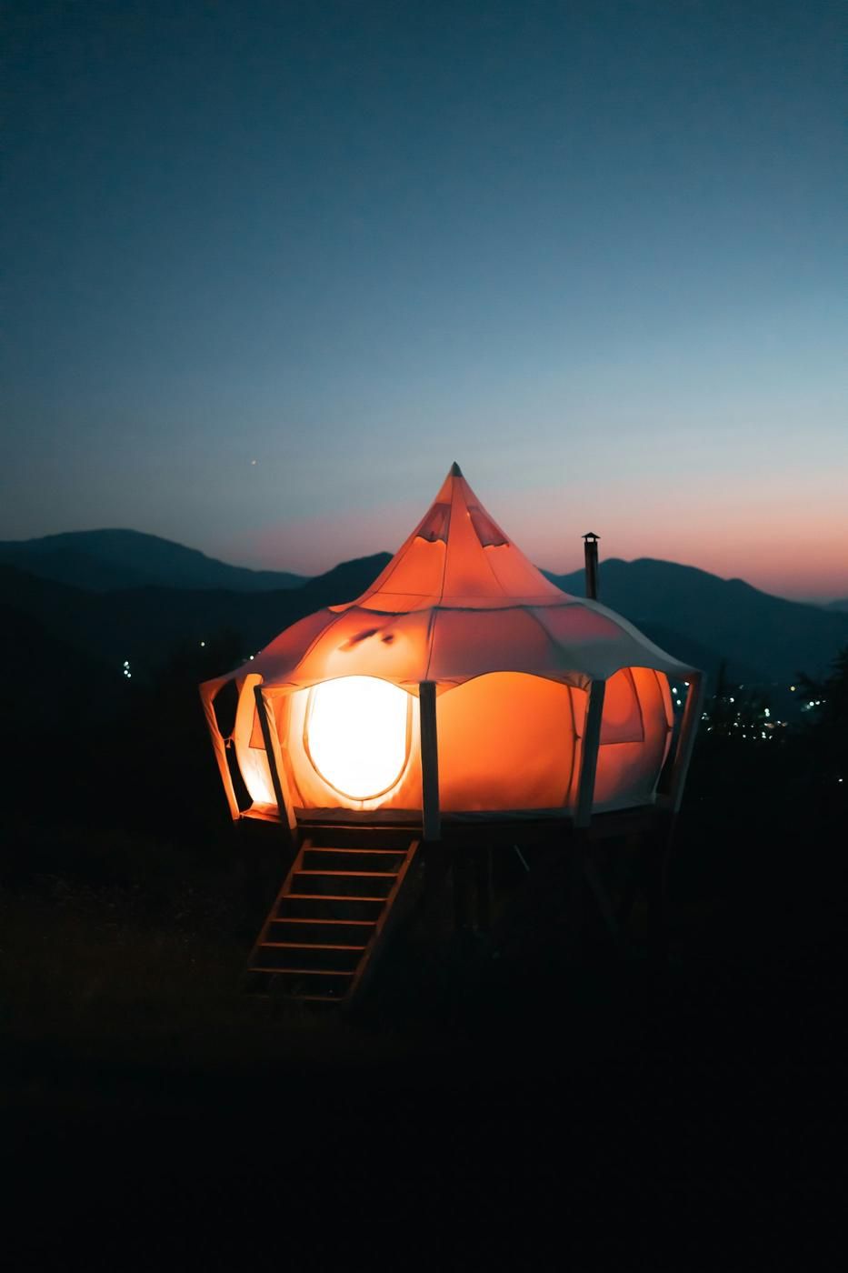 Tente de glamping illuminée de l'intérieur à la tombée de la nuit, devant des montagnes (photo Unsplash provisoire)