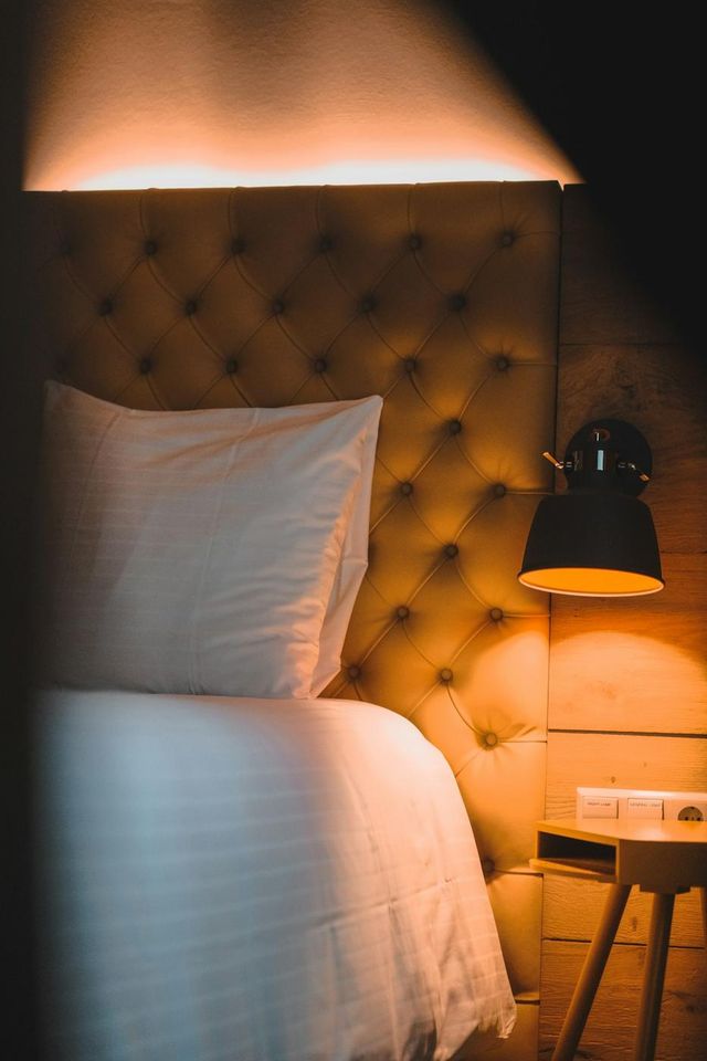 Lit d'hôtel aux draps blancs devant une tête de lit capitonnée, éclairée par une applique à la lumière chaude (photo Unsplash provisoire)
