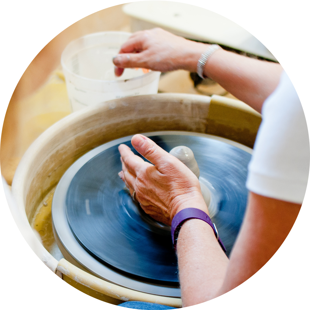Closeup of a pair of female arms working on a small clay formation spinning on a potter's wheel. The right hand is dipping into a plastic jar of water while the left hand is shaping the clay.