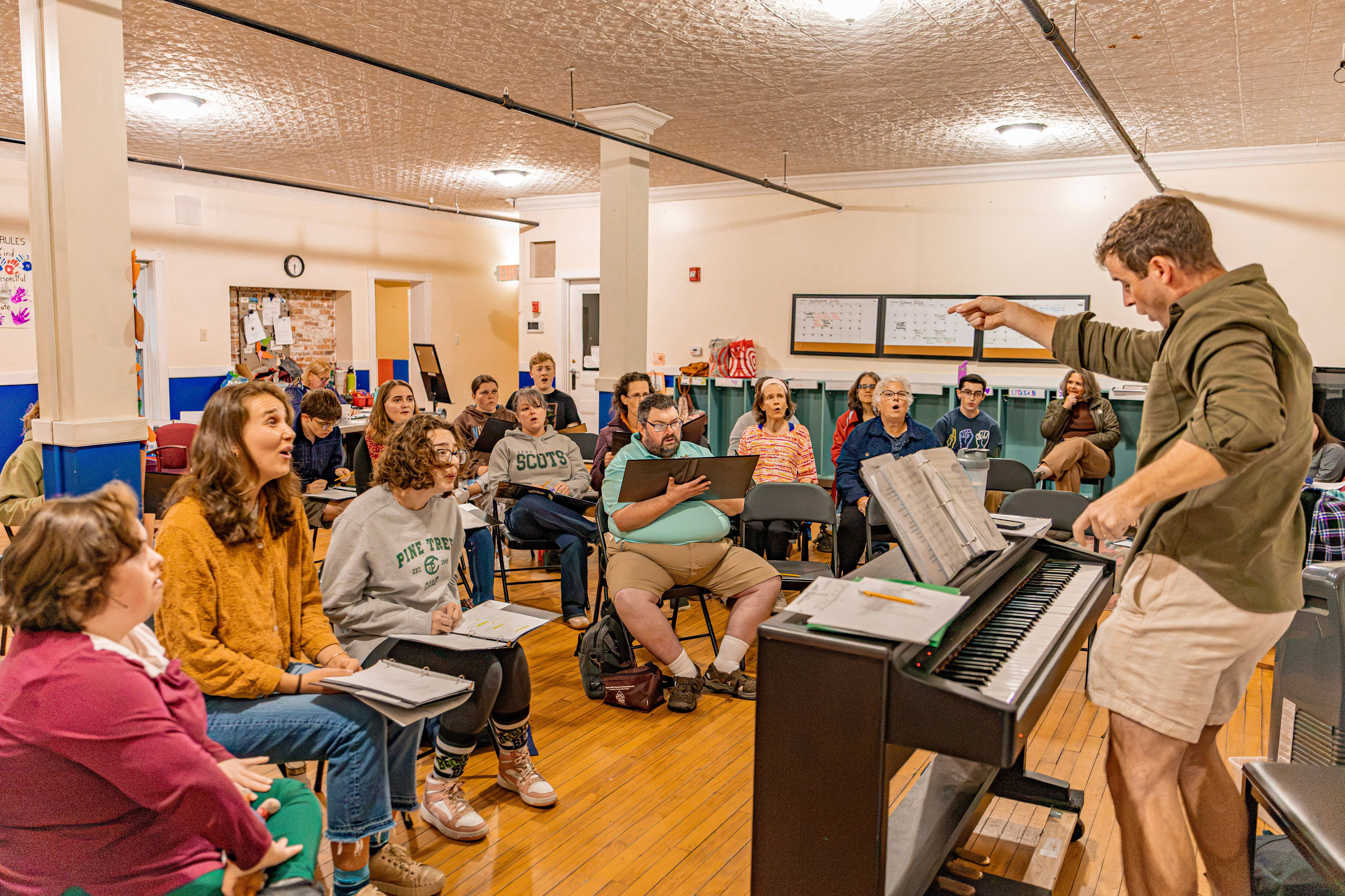 A theater director stands in front of a piano and directs a group of sitting performers