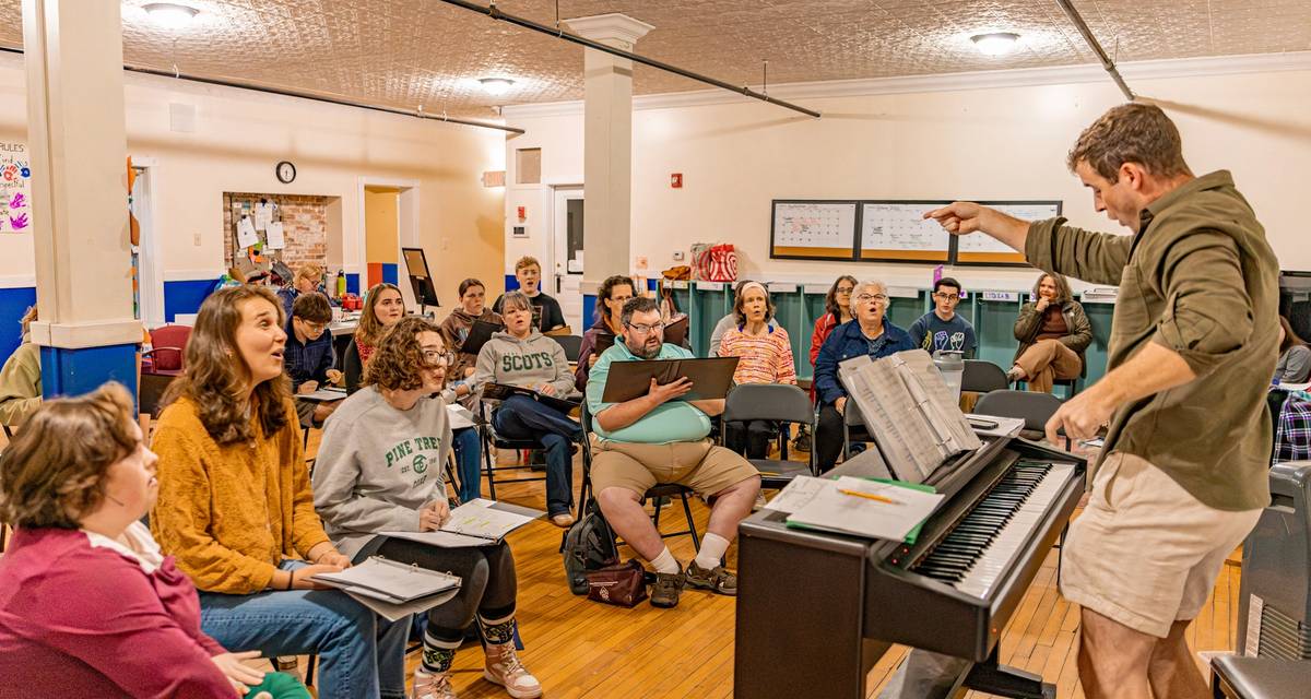 A theater director stands in front of a piano and directs a group of sitting performers
