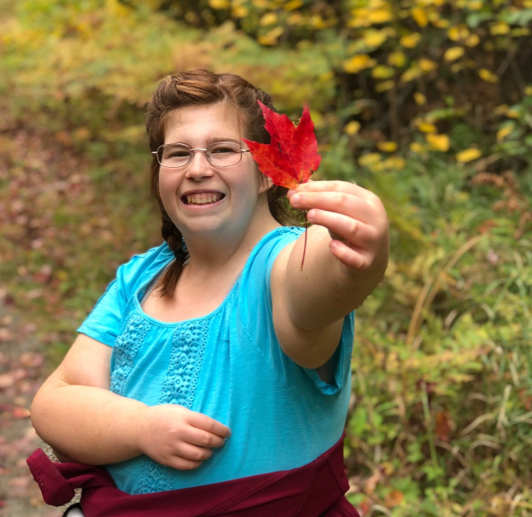 Young woman wearing a turquoise blouse holding a red maple leaf out to show us.