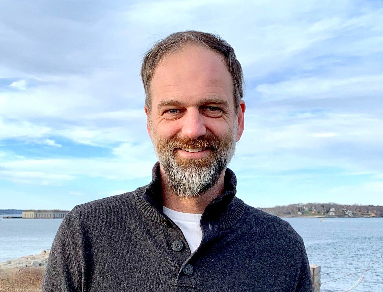 Nat May is standing in front of Casco Bay, with For Gorgeous, Peaks Island, blue sky and clouds behind him. He is smiling and looking at the camera.  
