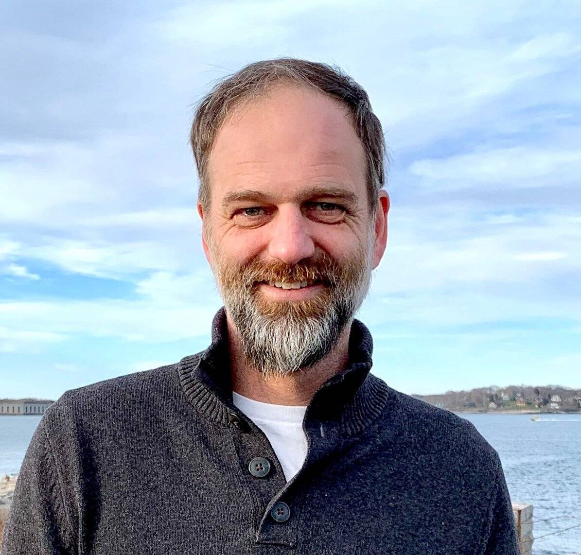 Nat stands in front of Casco Bay with a blue sky behind him. He is looking at the camera and smiling.