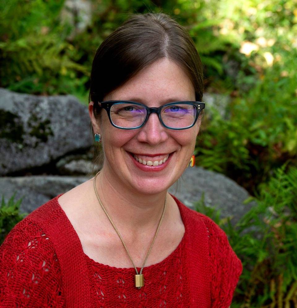 Sally sits in a outdoor space, smiling at the camera, wearing a red shirt