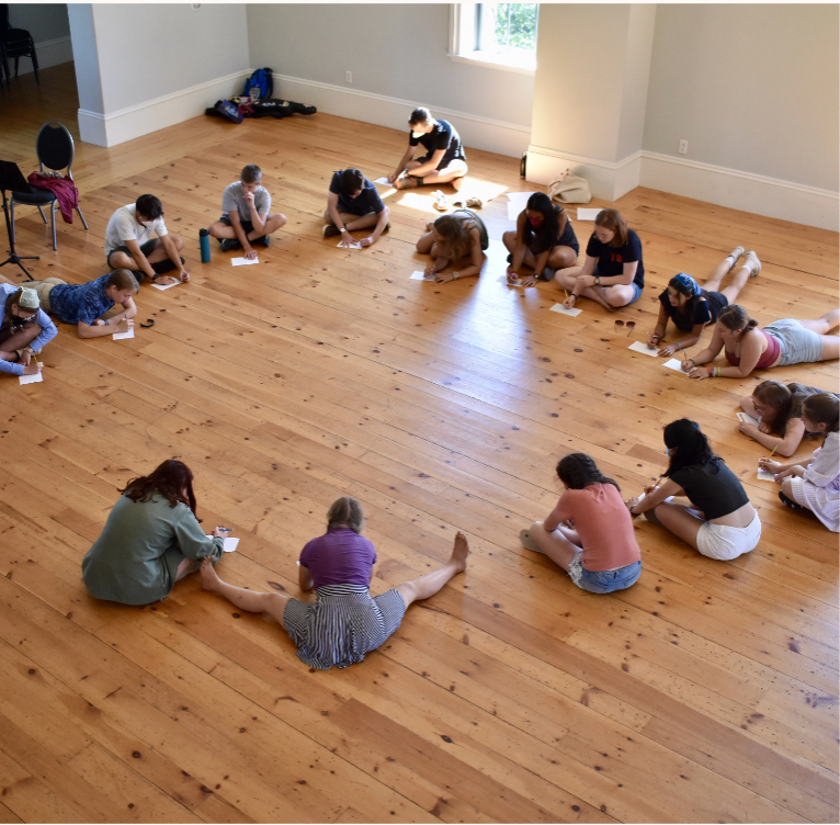 A group of school children sitting in a circle on a wooden floor writing on pieces of paper.