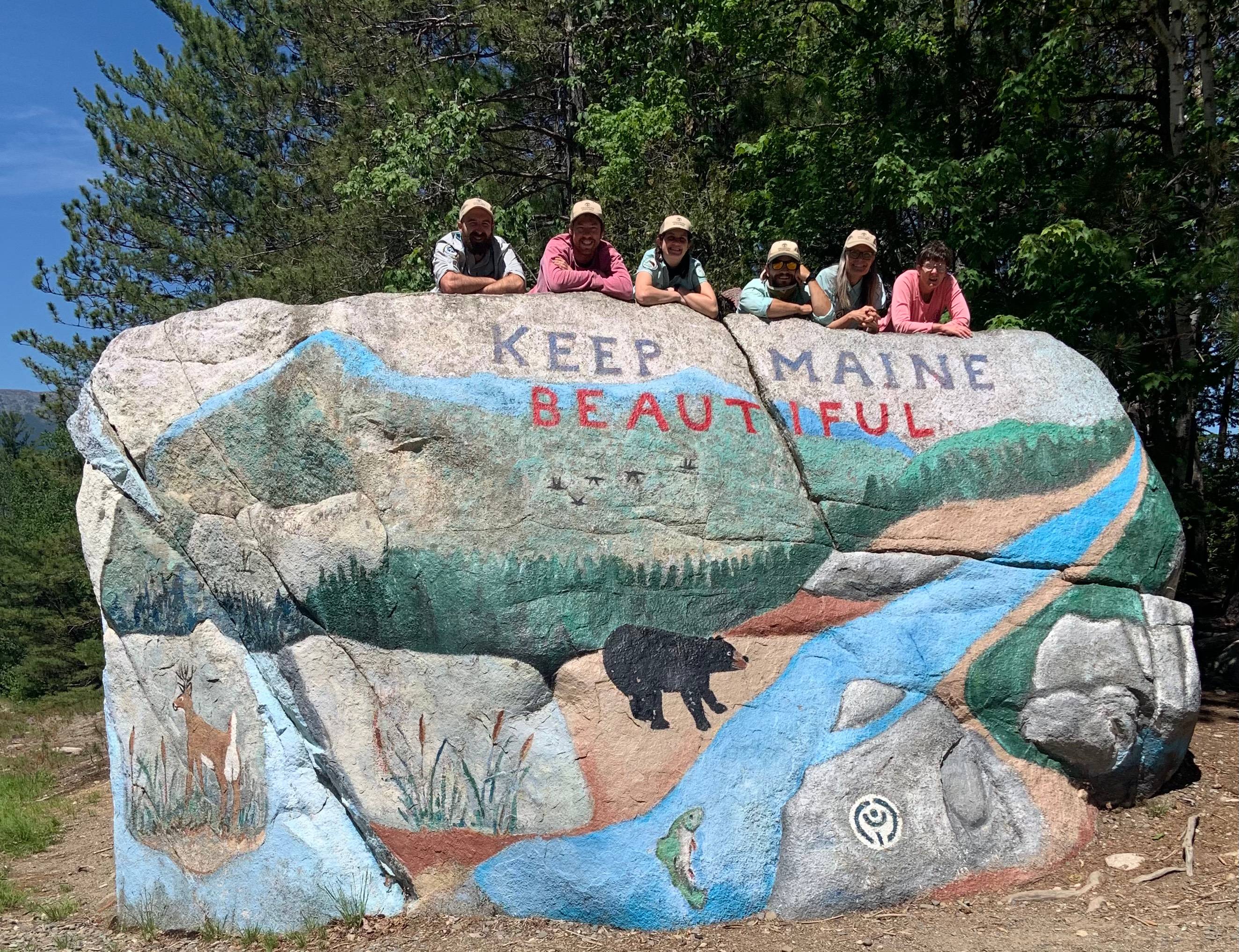 Six trail hikers lay atop a boulder featuring a mural of Maine
