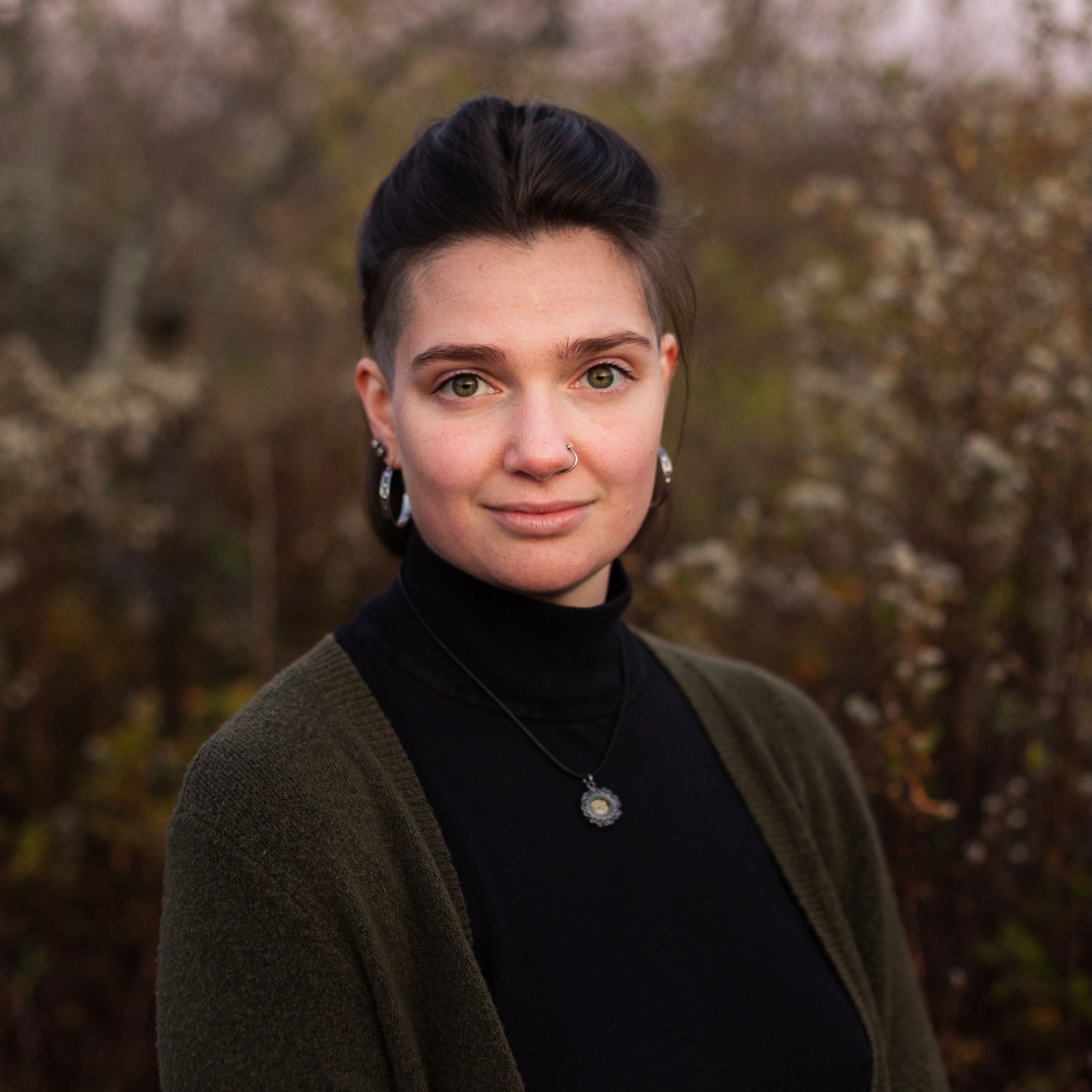 Holly is looking directly at the camera with a dlight smile. She is outside at Kettle Cove at dusk, with tall dune grasses in the background. 
