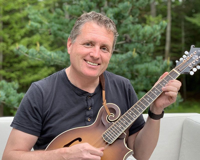 Fritz is sitting outside playing his mandolin. He is wearing a black t-shirt and smiling at the camera.