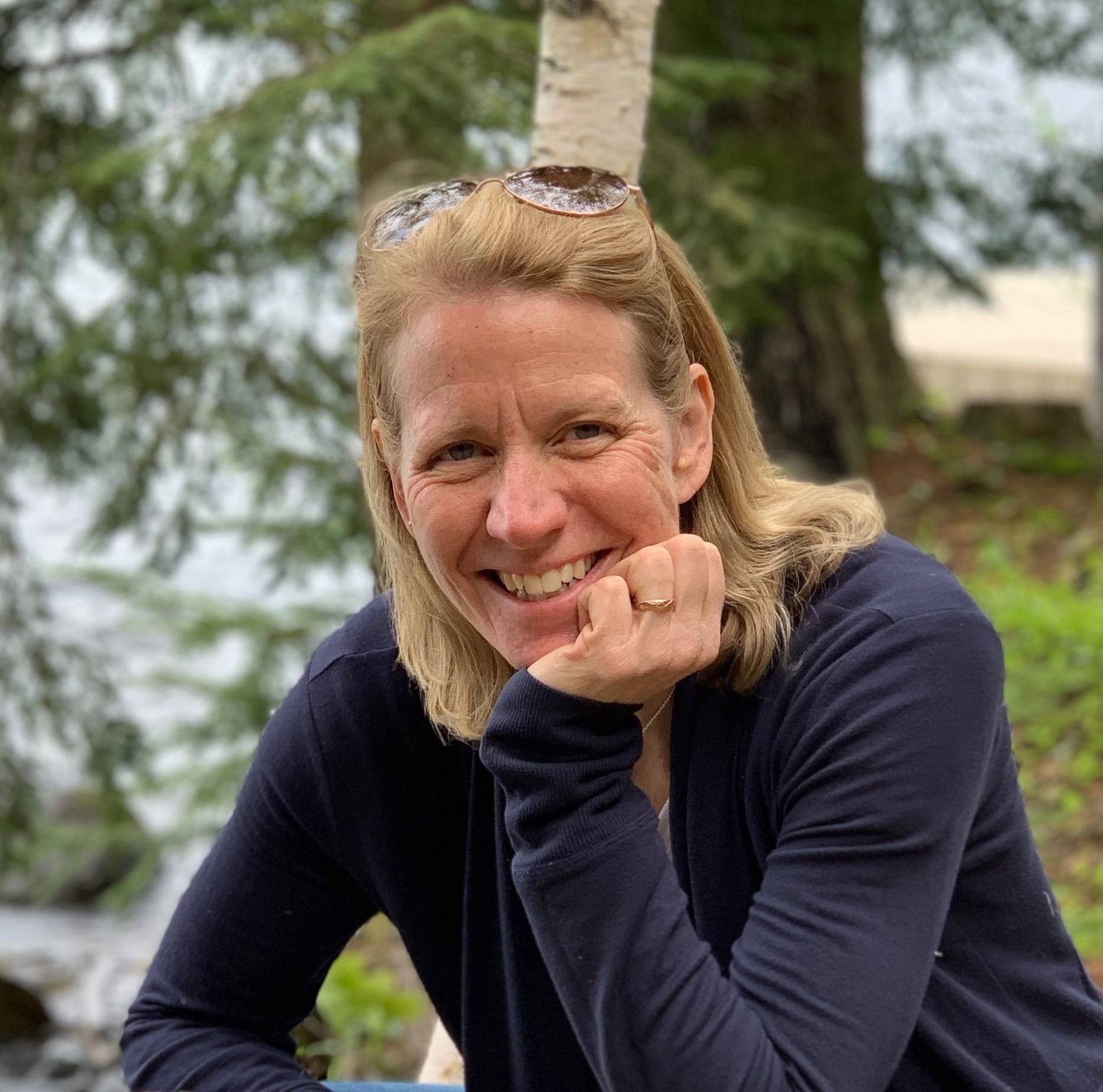 Susan sits outside and smiles at the camera, with her chin propped on her hand. 