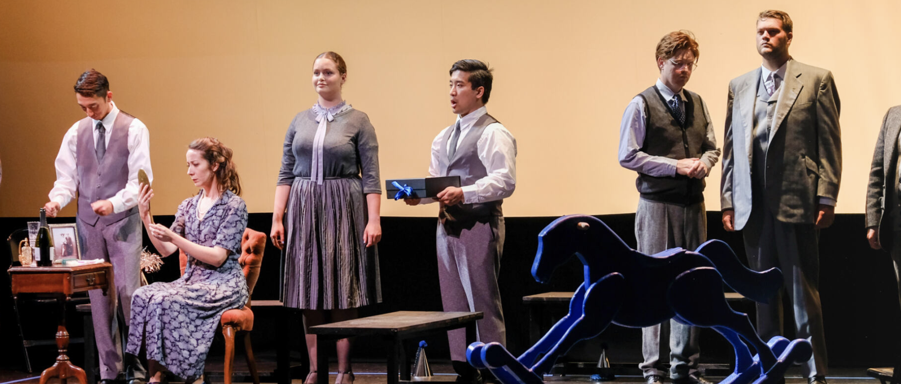 A group of people on stage in costume mostly grey and white. Most are standing, but one woman on the left is sitting. One man has his mouth open as if singing.