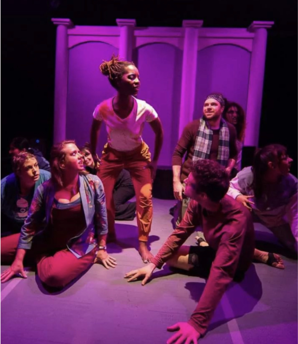 A scene from a theater production with seven actors in various seated positions surrounding a female actress standing with her hands on her thighs.