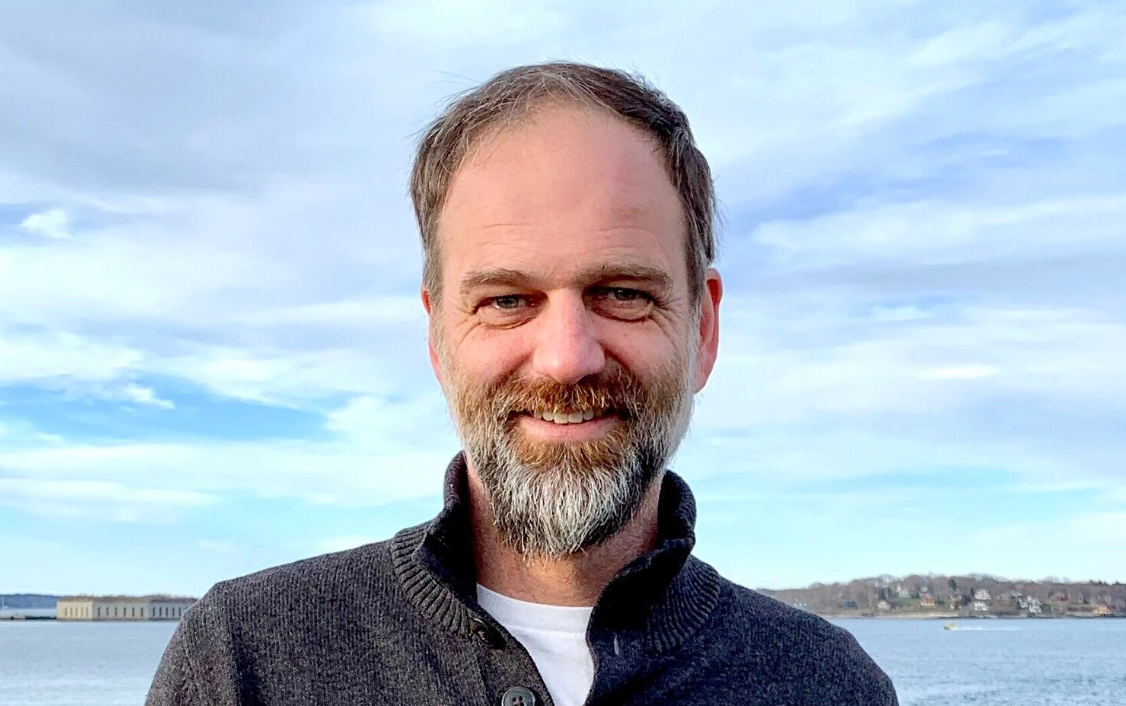 Nat May is standing in front of Casco Bay wearing a grey sweater and smiling at the camera. 