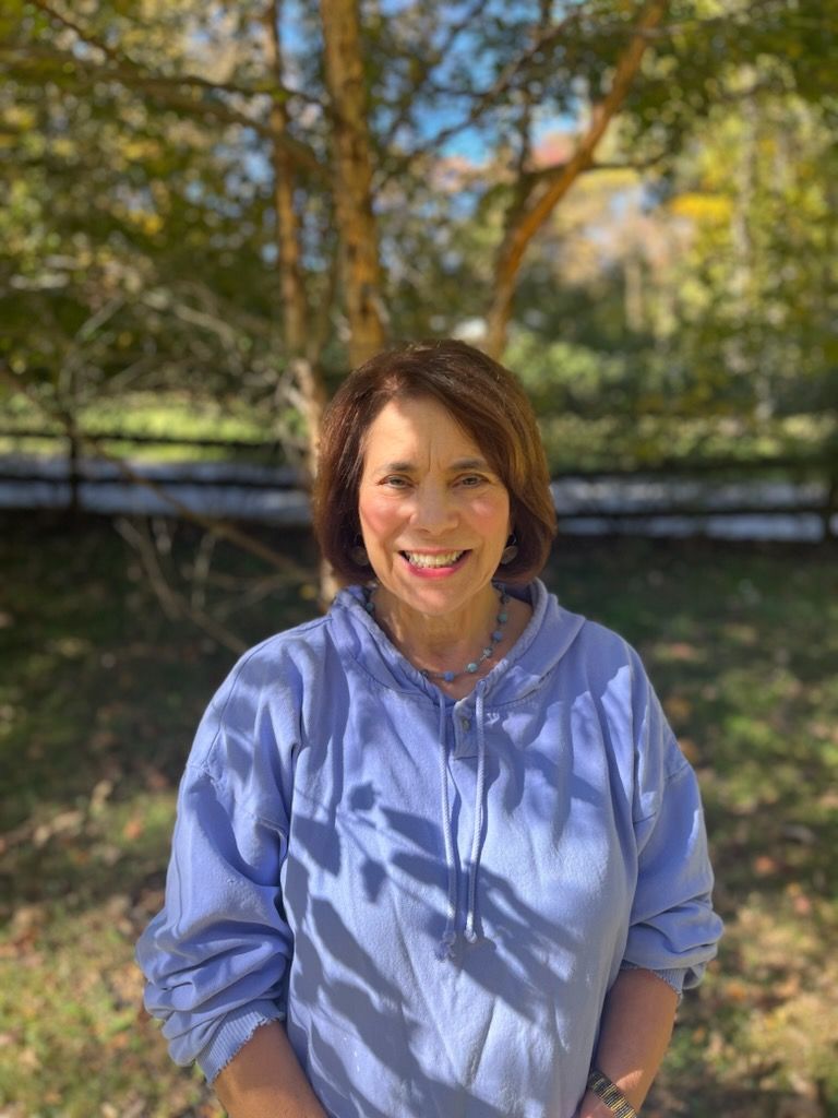 Diana Morris is standing outside in dappled shade, smiling at the camera. she is wearing a blue shirt and beaded necklace. 