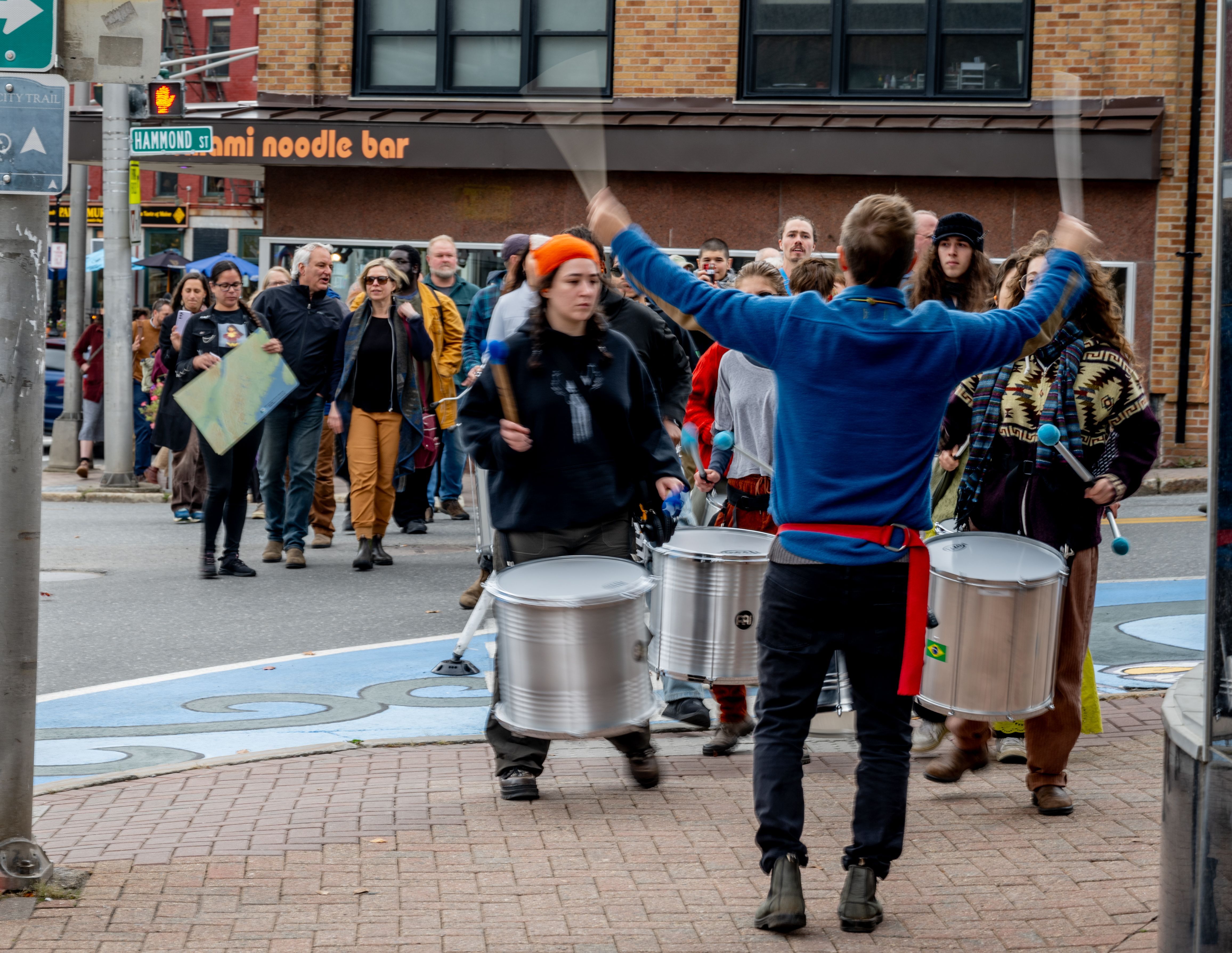 Three people with drums and a conductor march down a downtown road
