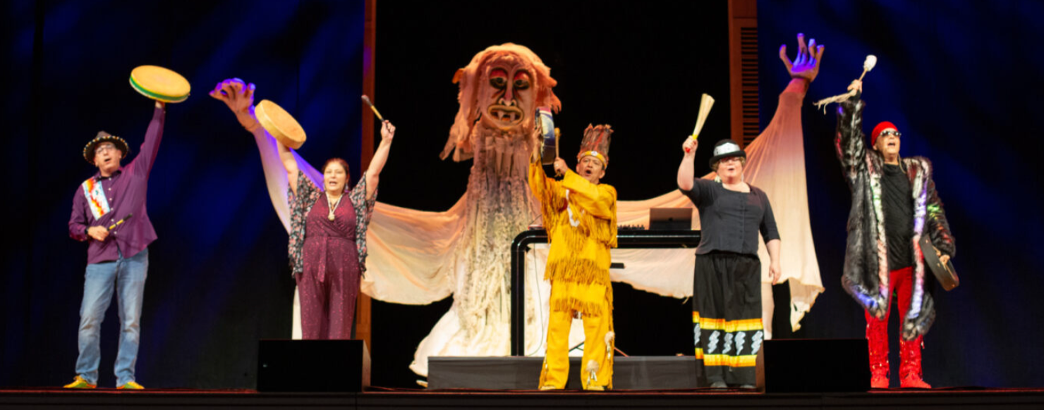 A scene from a theater production with five actors dressed in brightly colored clothes holding hand drums and mallets with a bigger than life puppet behind them.