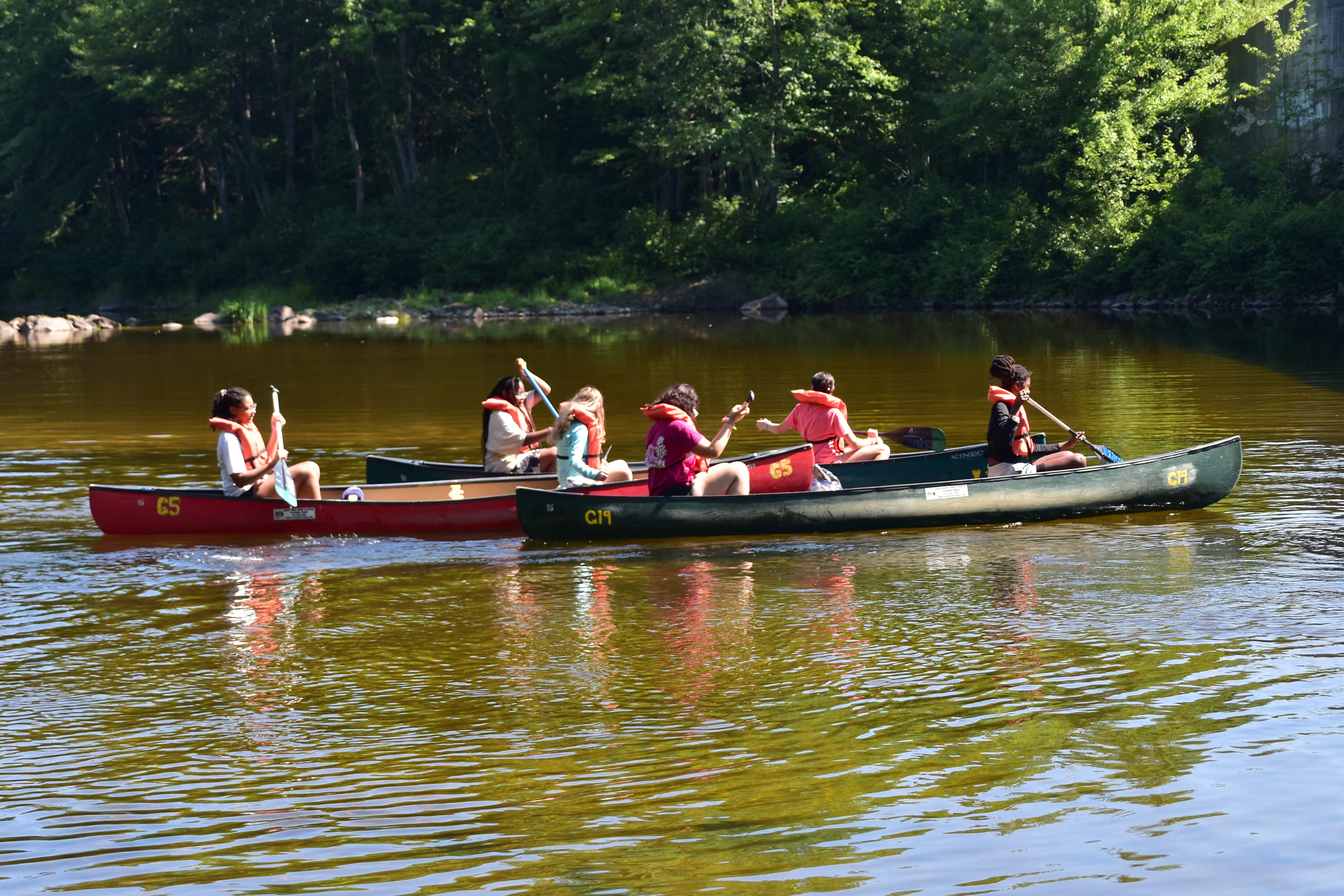 Multiple young people paddle canoes on a lake