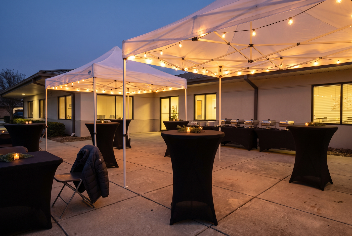 Office courtyard set for a holiday party at dusk, with white canopy tents, string lights, black spandex cocktail tables, and a buffet line of chafing dishes