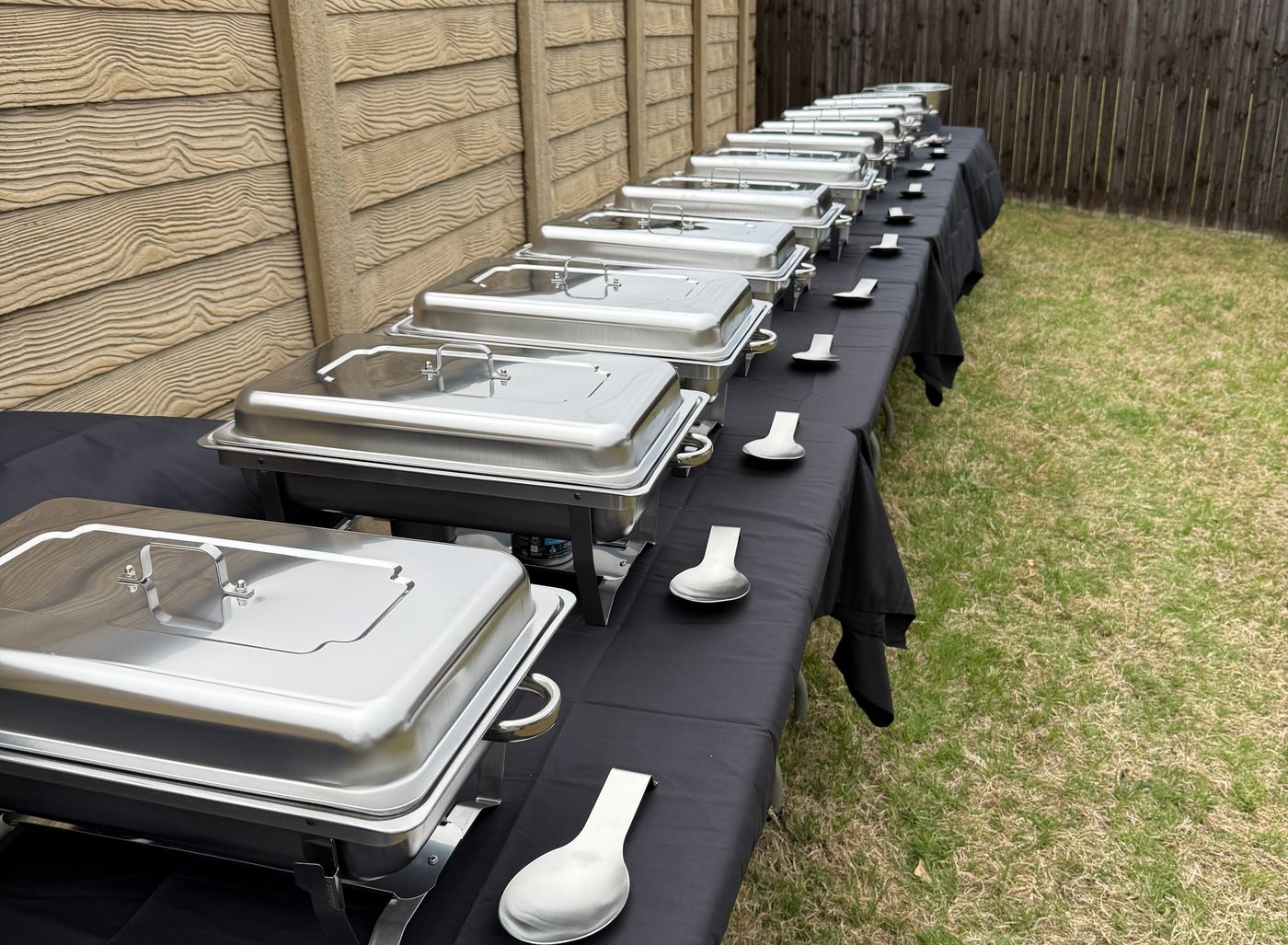 Buffet line of stainless steel chafing dishes on black-draped tables set up in a backyard