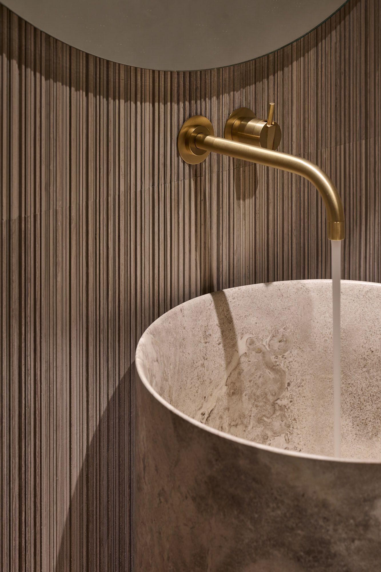 Detailaufnahme eines puristischen Waschplatzes mit goldener Armatur, vertikaler Strukturwand und zylindrischem Natursteinbecken für ein warmes, modern elegantes Ambiente.