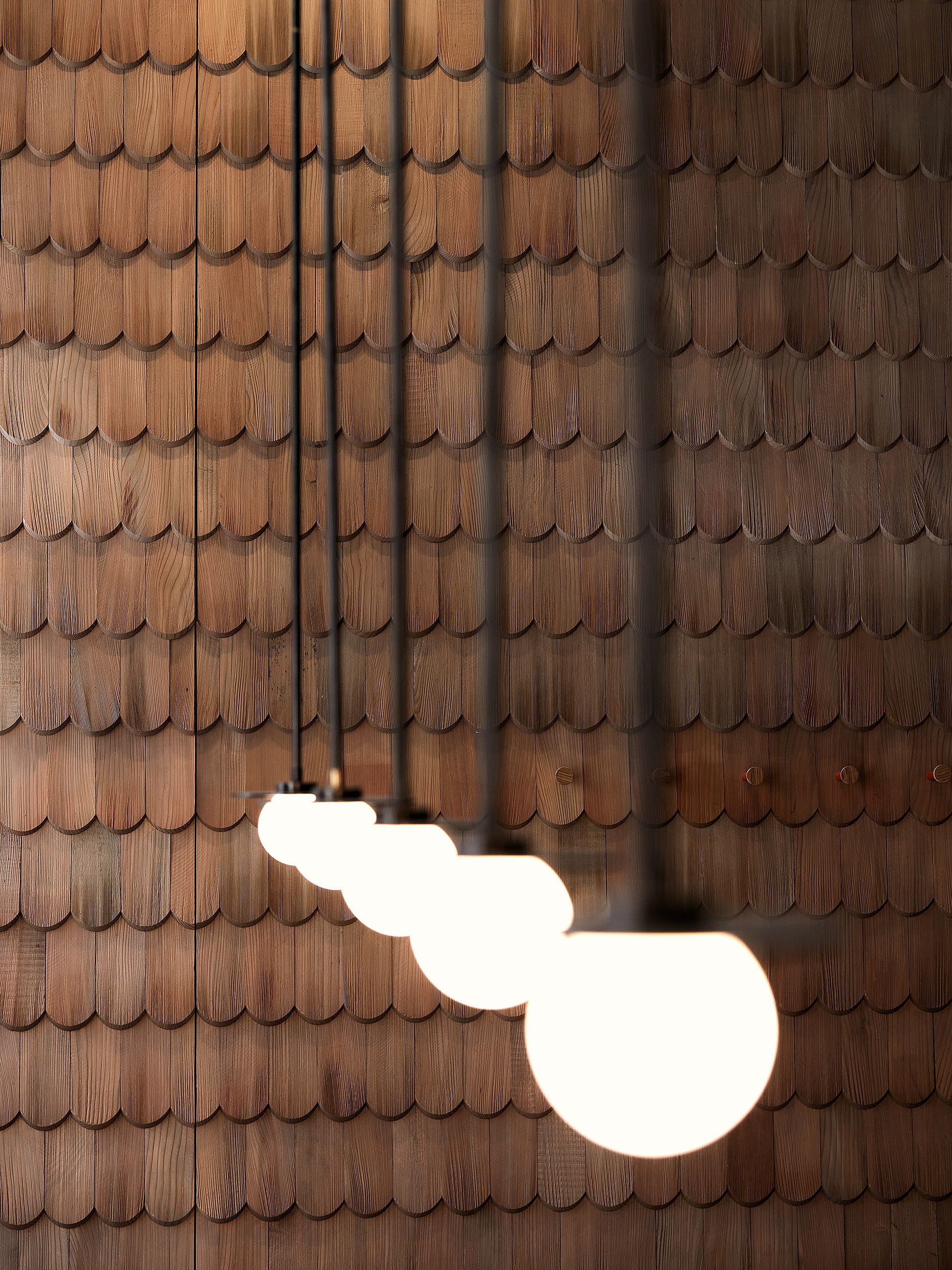 Die rhythmisch gehängten Leuchten vor einer Wand aus Holzschindeln schaffen im Benediktiner Weissbräuhaus eine warme, atmosphärische Lichtstimmung im Stil von Stephanie Thatenhorst.