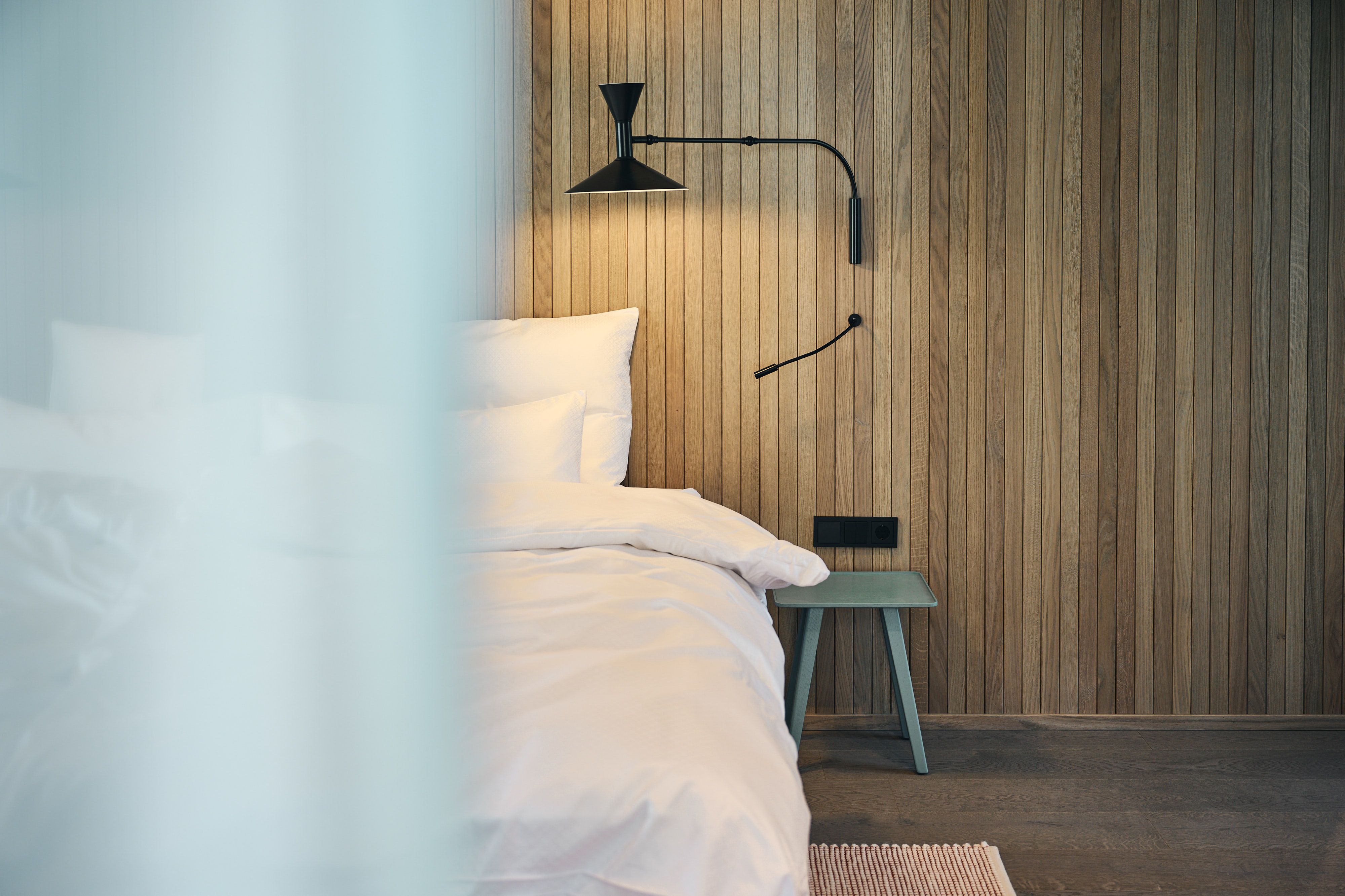 Das Bild zeigt ein von Stephanie Thatenhorst gestaltetes Ullrhaus-Schlafzimmer, geprägt von einer ruhigen Holzvertäfelung und einer minimalistischen schwarzen Wandleuchte.