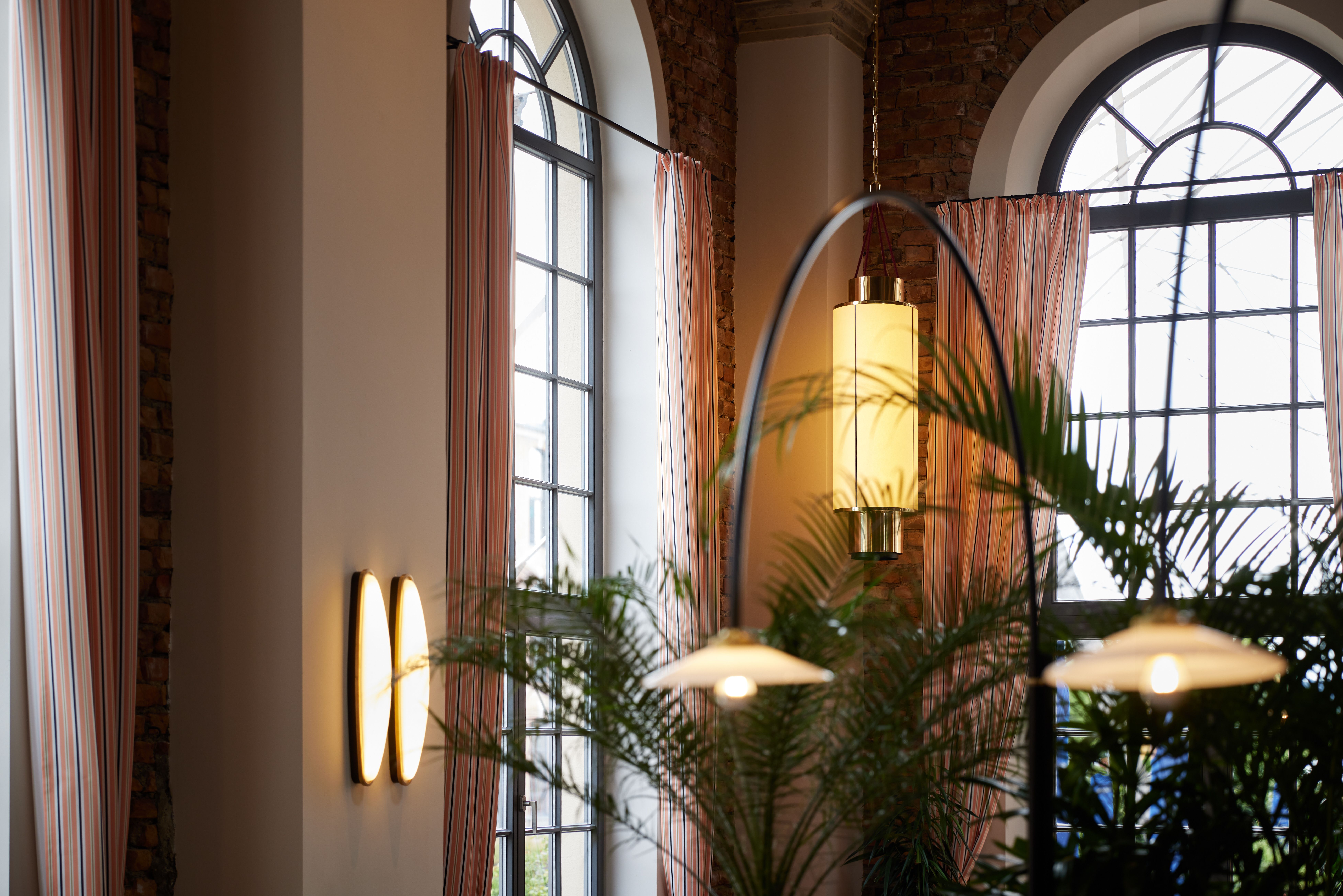 Das raumhohe Rundbogenfenster-Setting mit gestreifter Drapierung, Art-Deco-Wandleuchte und begrüntem Zonierungskonzept wurde im Turbinenhaus Kolbermoor – Brasserie von Stephanie Thatenhorst designt.