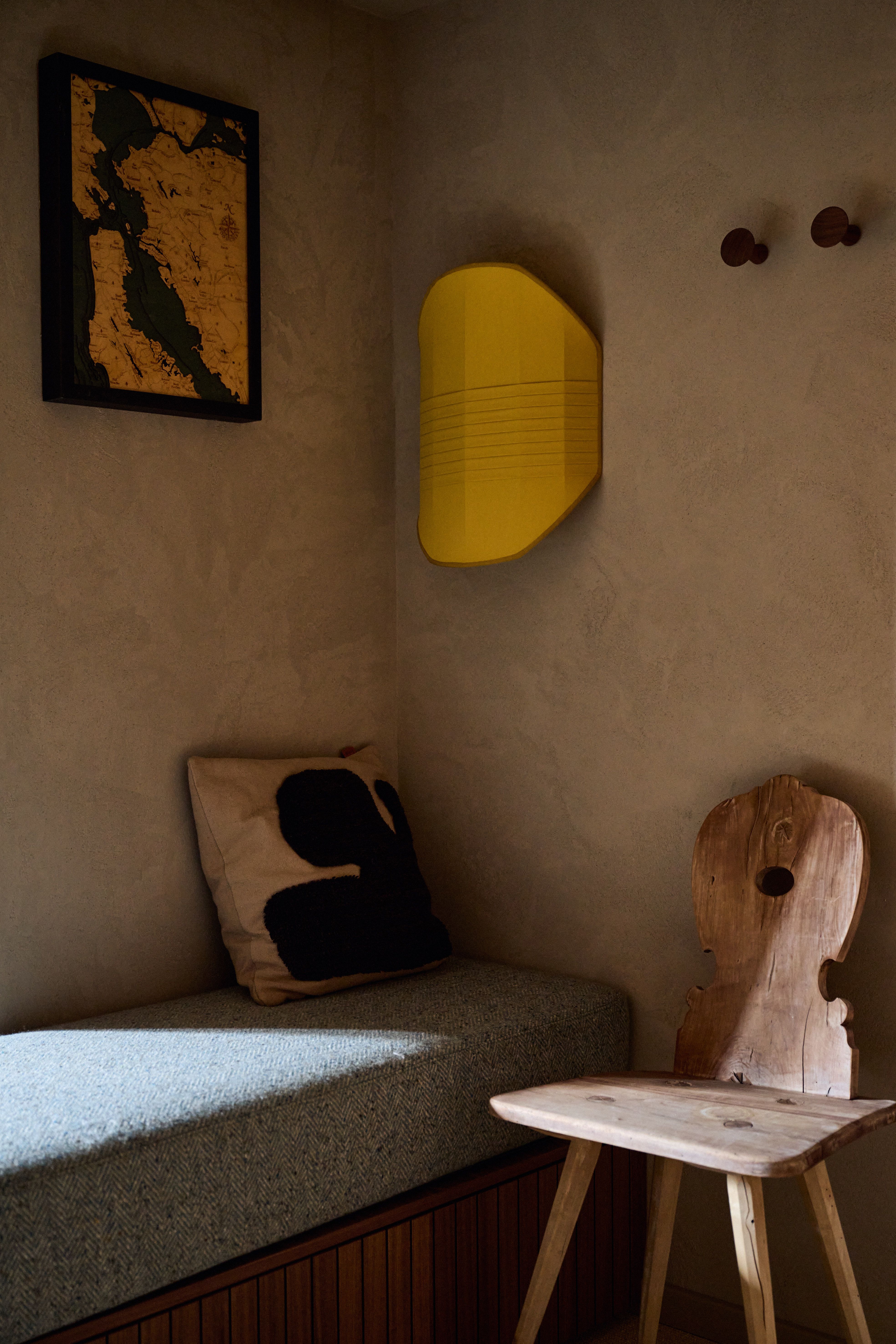 Gemütliche Sitzecke mit rustikalem Holzstuhl, grafischem Kissen und gelber Wandleuchte im spannenden Kontrast zur natürlichen Putzstruktur.