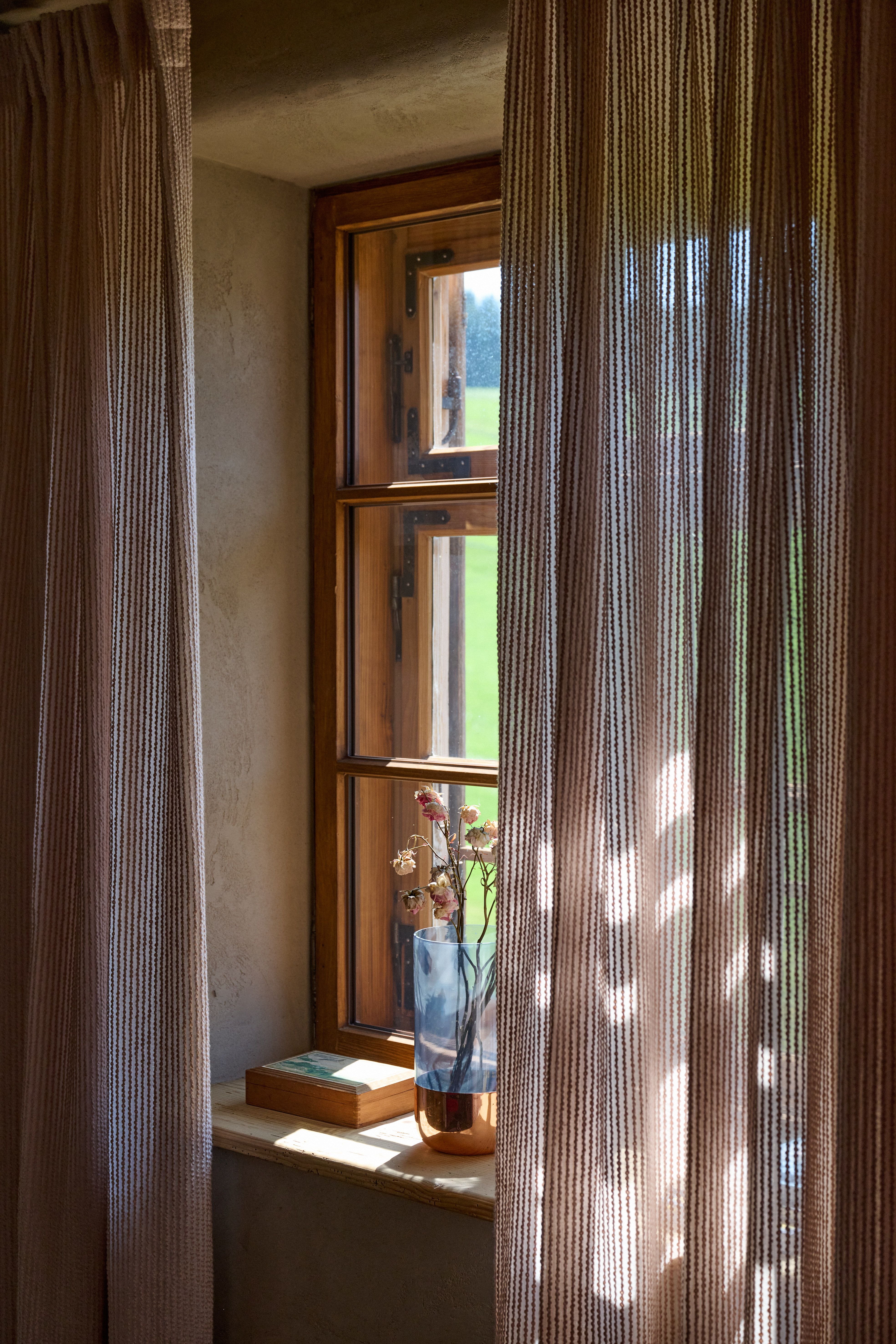 Designed by Stephanie Thatenhorst, entfaltet dieses intime Fensterarrangement weiches Tageslicht, texturierte Vorhangtransparenz und warmes Holz zu einer stillen, atmosphärischen Landhauspoesie.