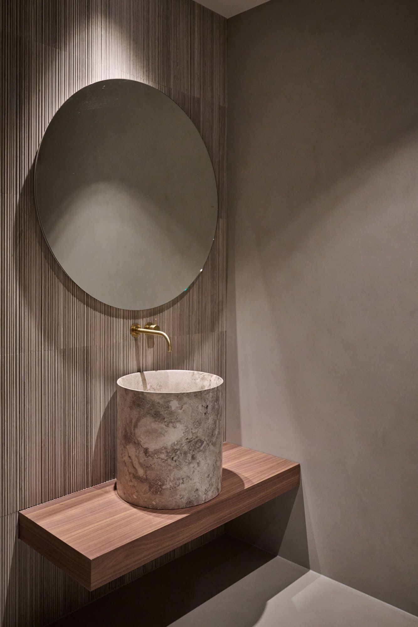 Puristischer Waschplatz mit rundem Spiegel, vertikaler Holzstruktur und zylindrischem Natursteinbecken für ein ruhiges, modern elegantes Ambiente