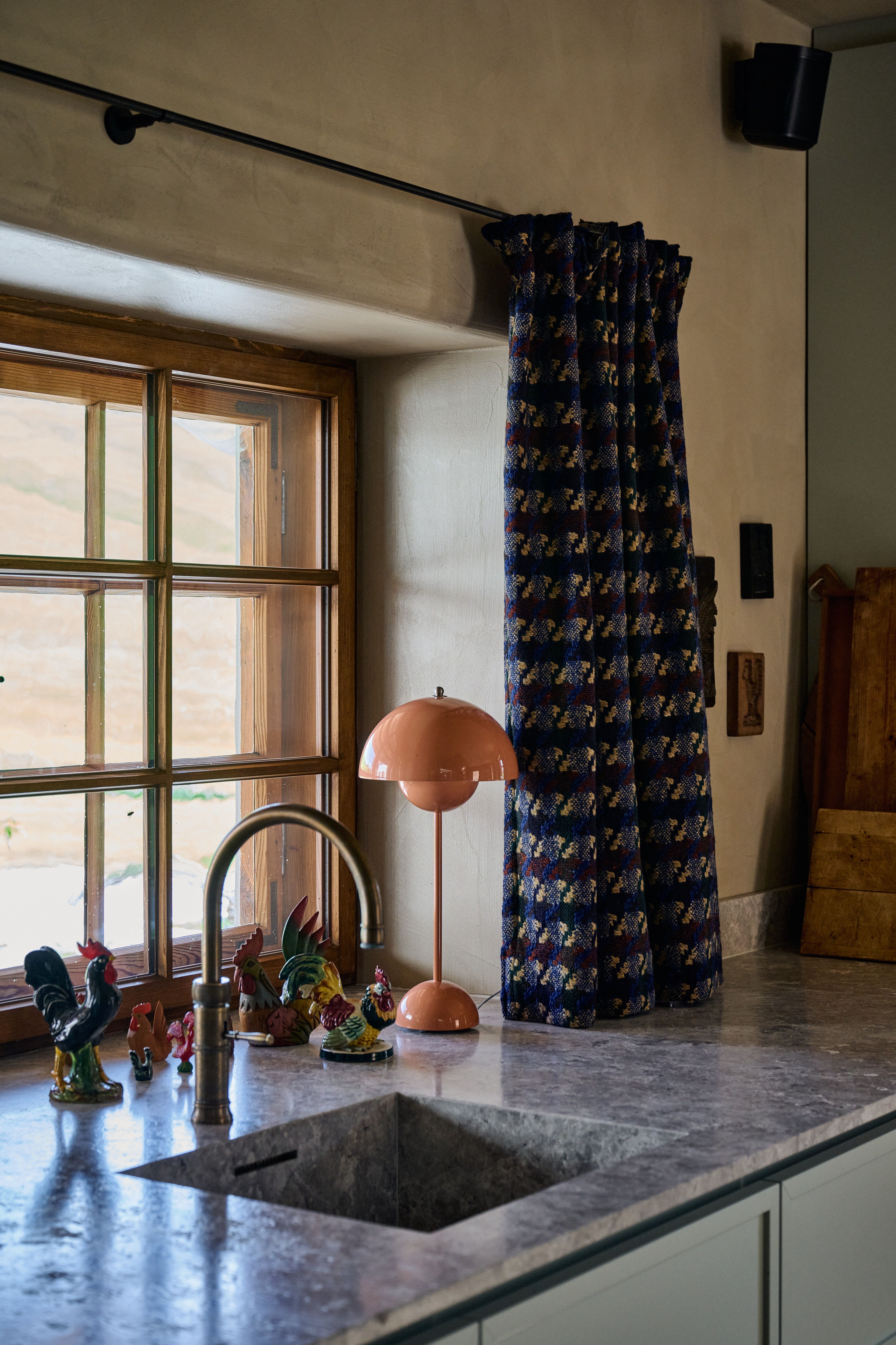 Designed by Stephanie Thatenhorst, verbindet dieses Küchenfensterarrangement strukturierte Steinoberfläche, grafisch gemusterte Vorhänge und sanft getöntes Licht zu einer warm akzentuierten, nostalgisch-modernen Landhauskulisse.