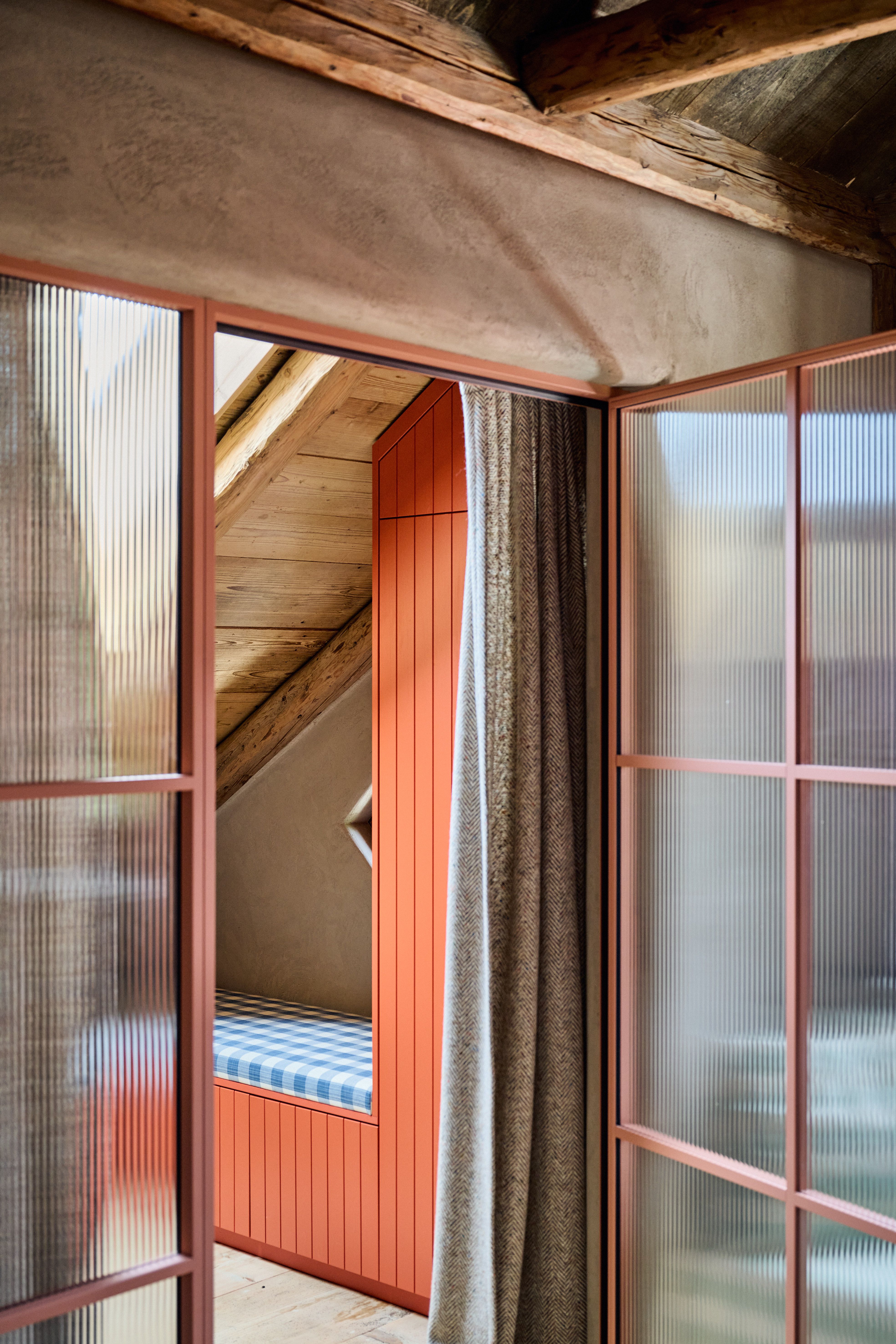 Raumtrennung mit roséfarbenen Profilglasschiebetüren und sichtbaren Holzbalken, kombiniert mit textiler Sitznische im Chalet-Look.