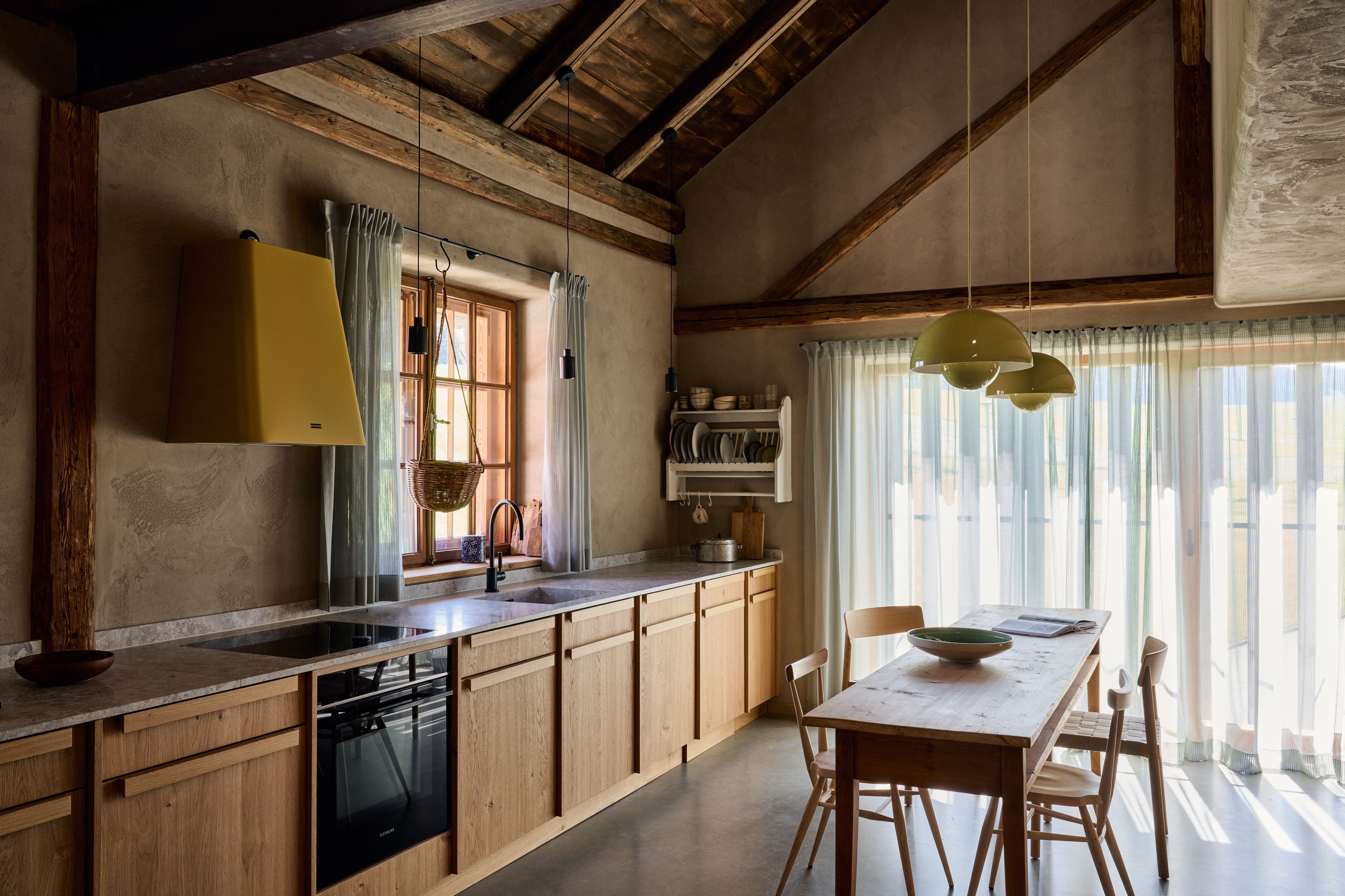 Offene Landhausküche mit massivem Holzausbau, sanft geglättetem Putz, gelber Dunstabzugshaube und großflächiger Fensterfront mit Lichtvorhängen.
