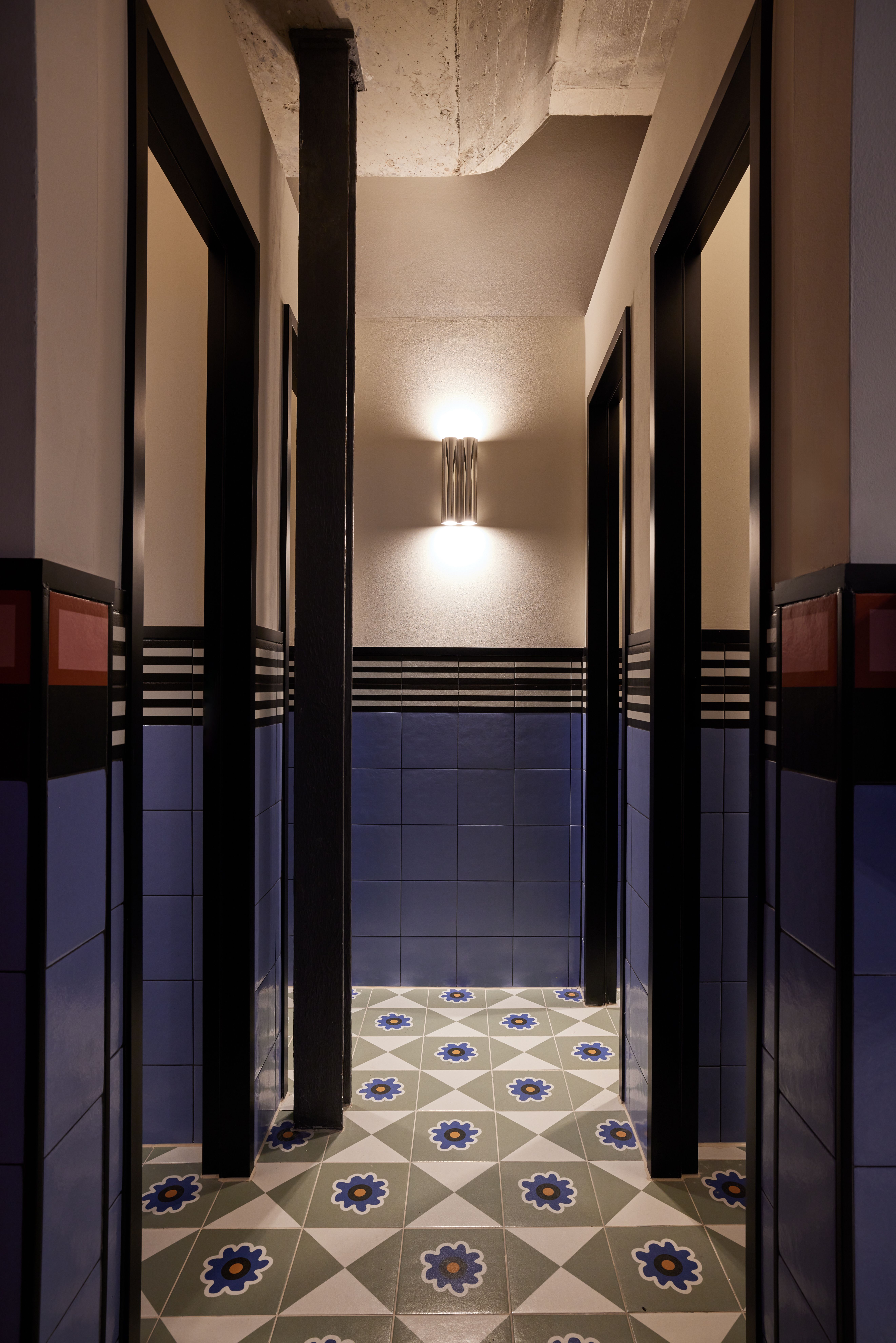 Die sanitäre Kabinenflucht mit grafischer Bordüre, keramischer Wandverfliesung und Boutique-Lichtobjekten wurde im Turbinenhaus Kolbermoor – Brasserie von Stephanie Thatenhorst designt.