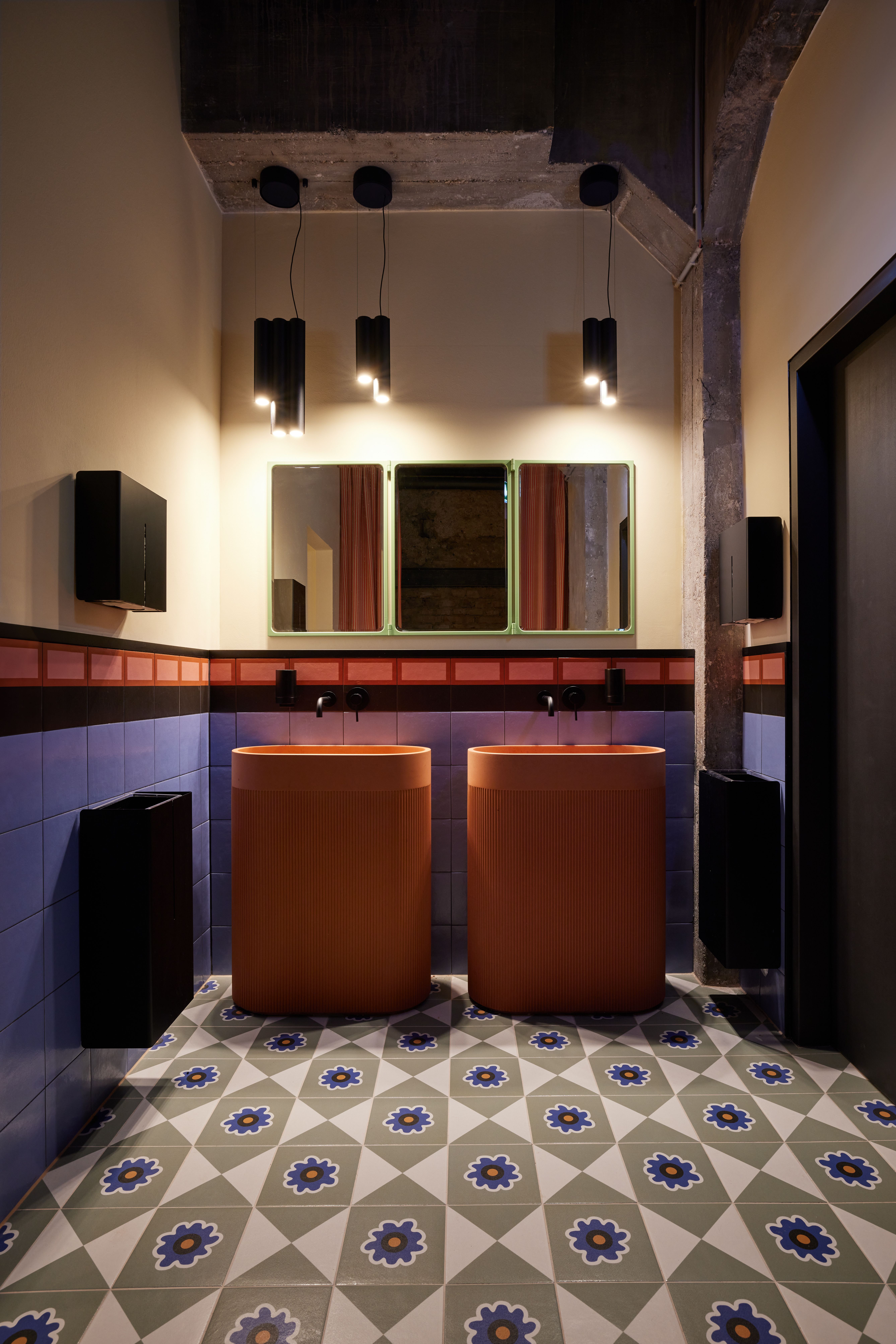 Der zweifarbige Waschraum mit freistehenden Terrakotta-Waschinseln, geometrischer Feinsteinzeug-Bodenfliese und industriellen Deckendetails wurde im Turbinenhaus Kolbermoor – Brasserie von Stephanie Thatenhorst designt.