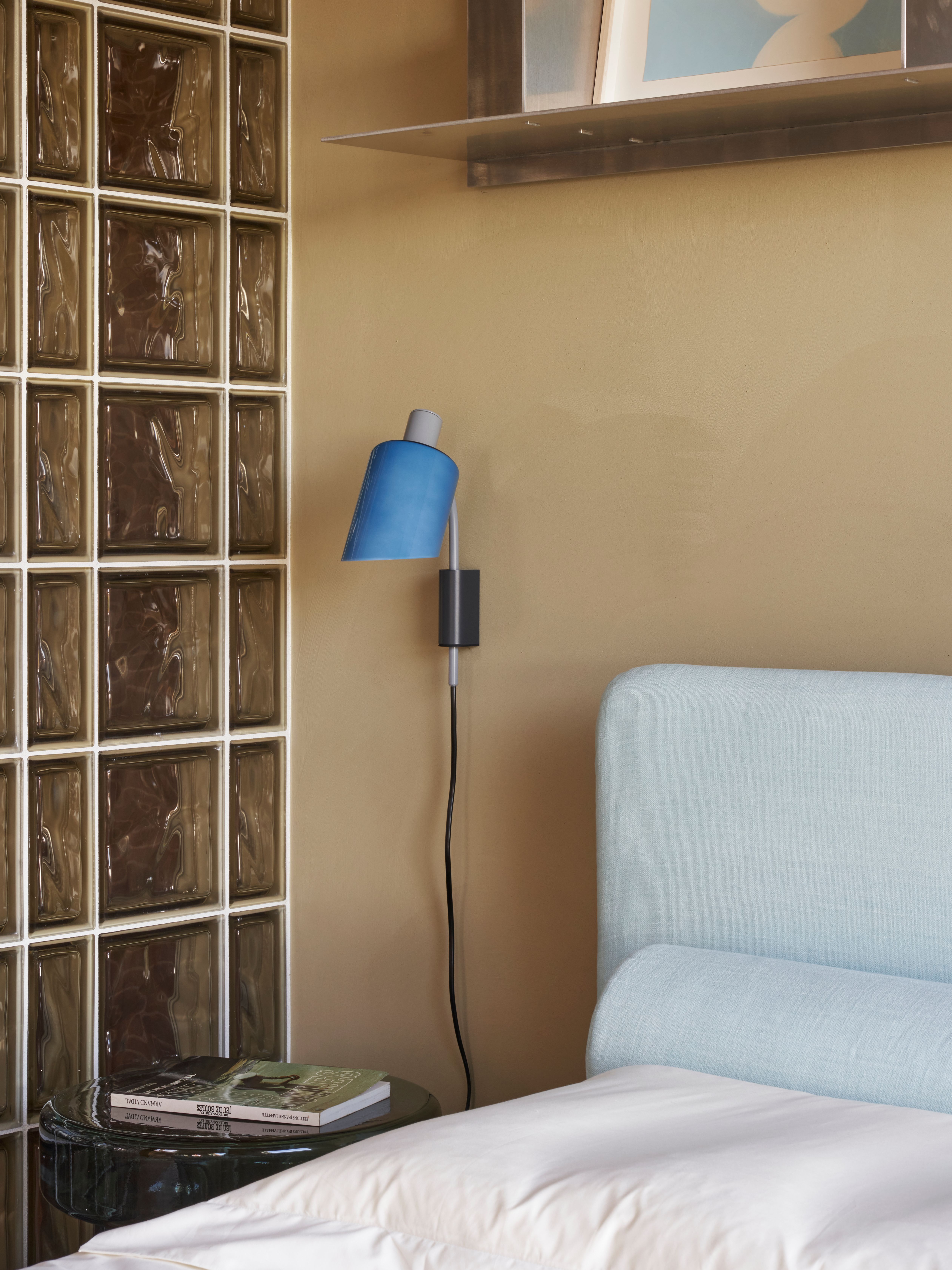 Die ruhige Schlafzimmerszene mit Glasbausteinwand, filigraner Ablage und ikonischer blauer Wandleuchte wurde von Stephanie Thatenhorst AD New Perspectives designt.