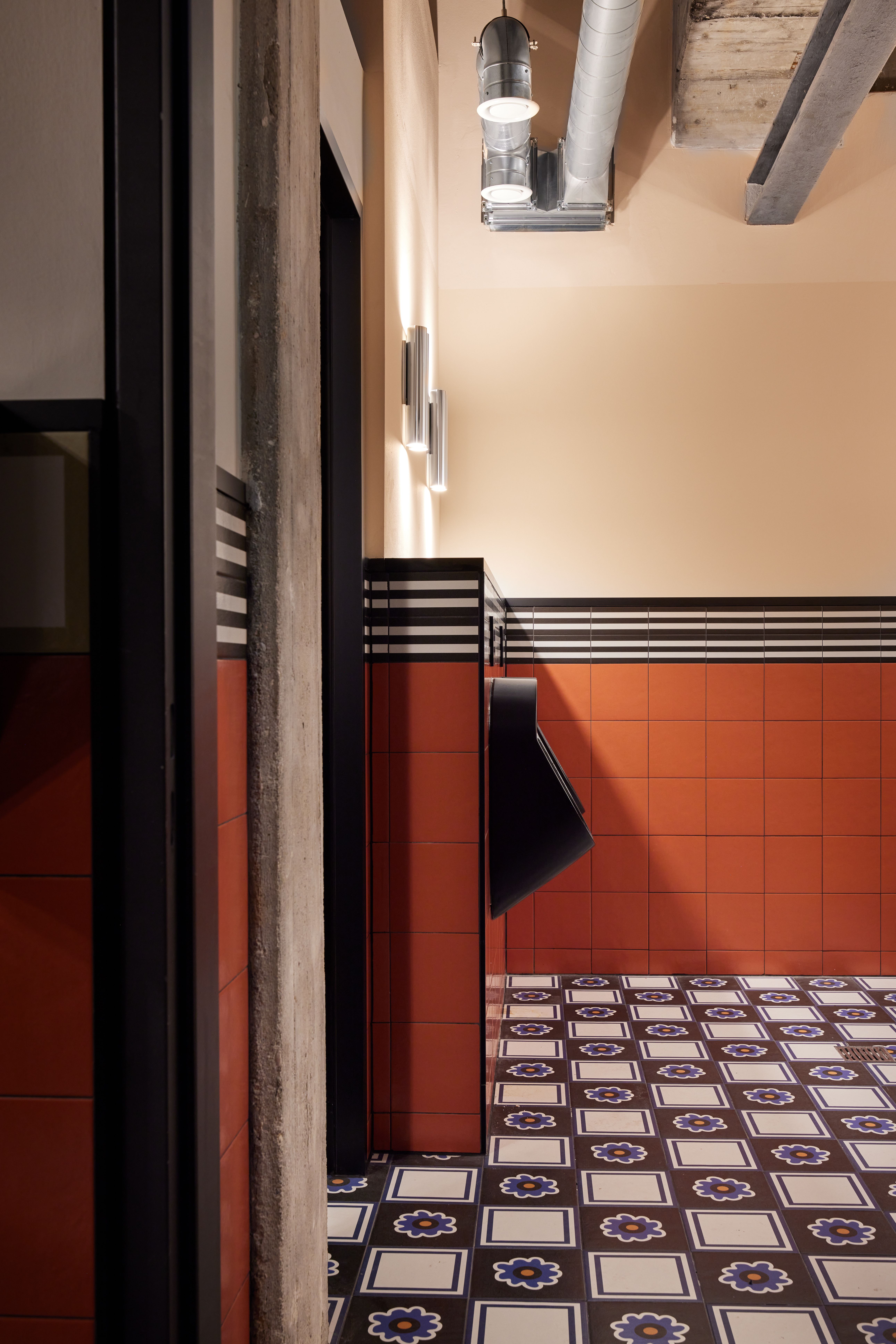 Der Waschraum mit Ziegelorange-Wandkacheln, architekturbetonter Sichtbeton-Stütze und floraler Zementmosaikplatte wurde im Turbinenhaus Kolbermoor – Brasserie von Stephanie Thatenhorst designt.