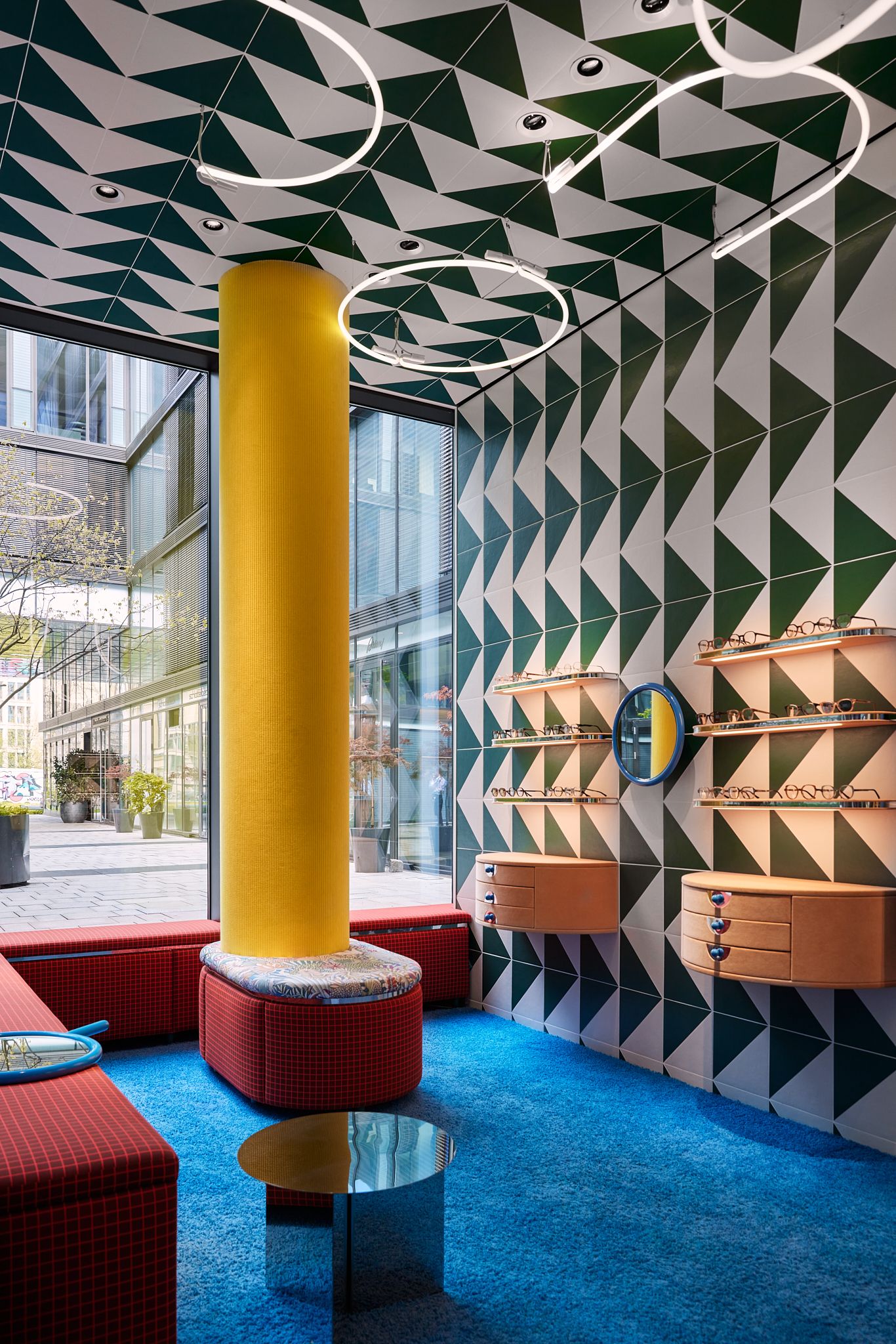 Die farbstarke Raumszene mit gelber Säule, geometrischer Wandgestaltung und individueller Möblierung demonstriert das lebendige Interior-Konzept, das Stephanie Thatenhorst für Rookies – Eyewear for Kids entwickelt hat.